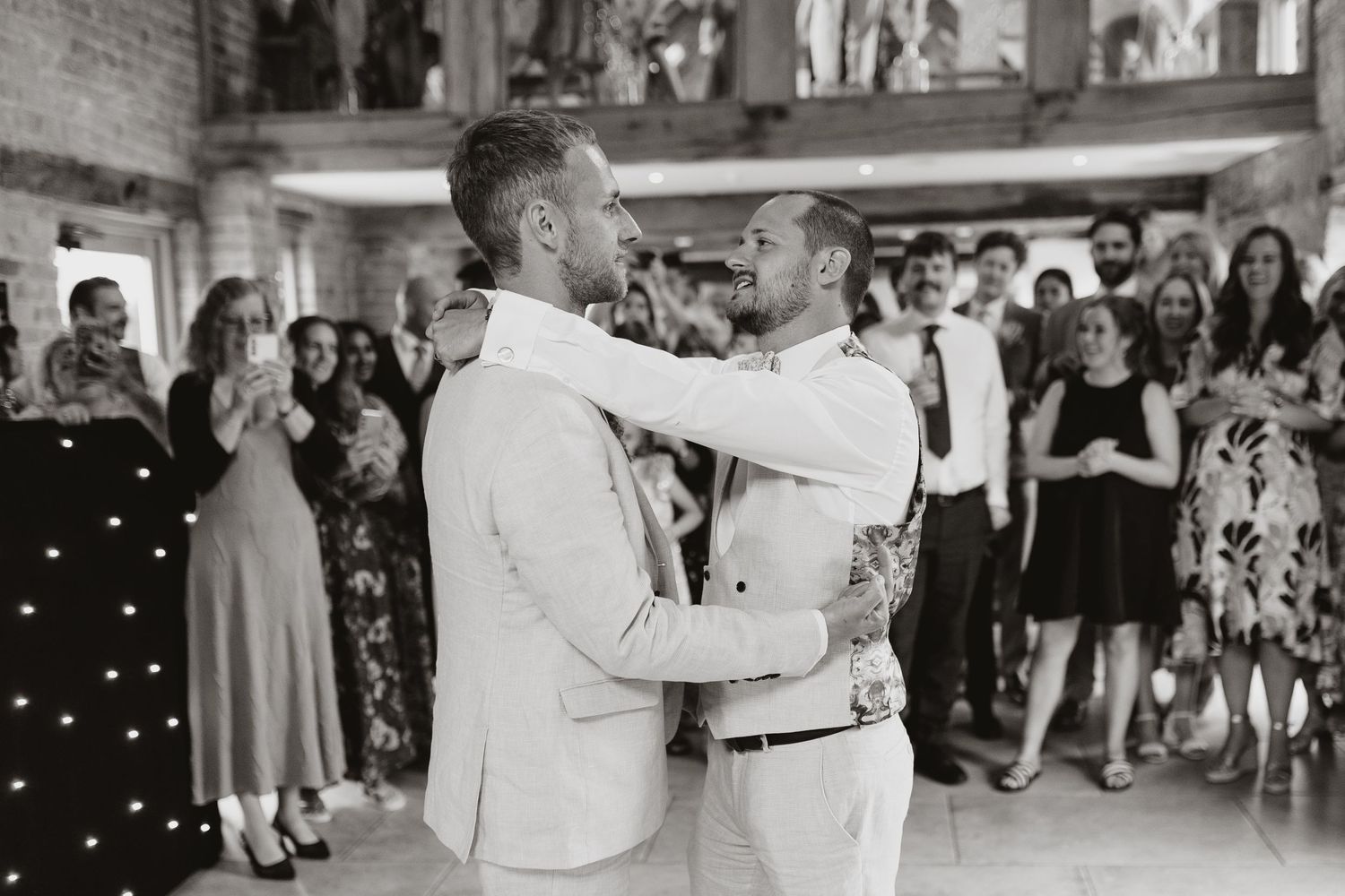 Two grooms share their first dance at a wedding reception, embraced together while guests applaud in the background.