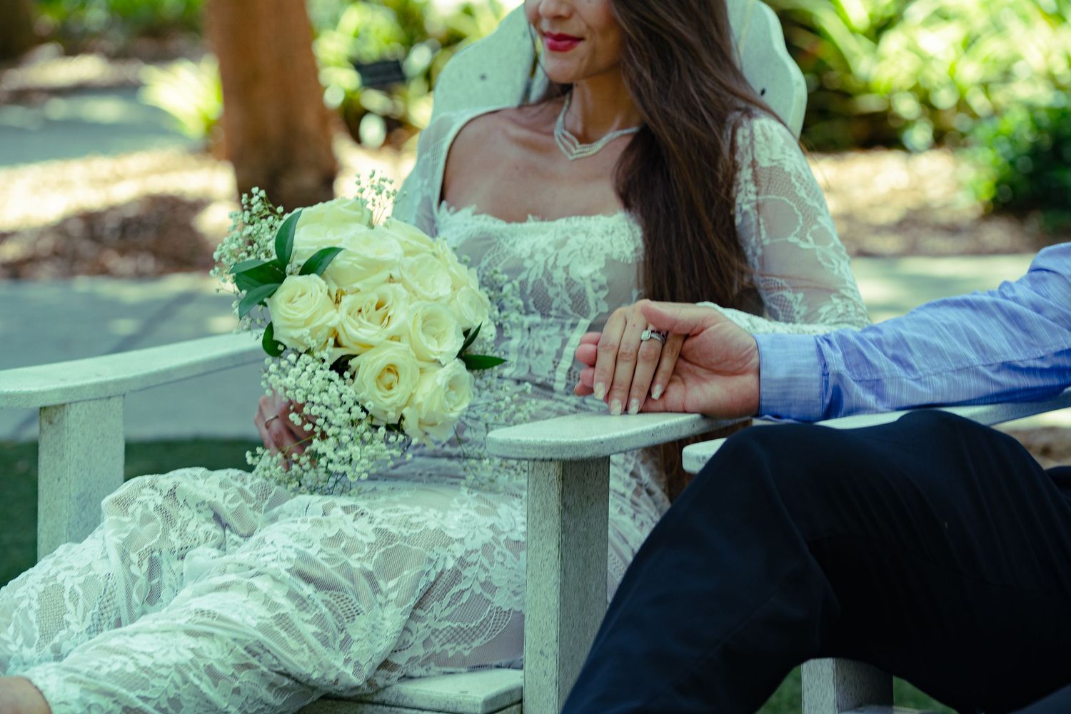 Bride in white lace gown holding yellow roses bouquet sits beside groom in blue shirt on garden bench.