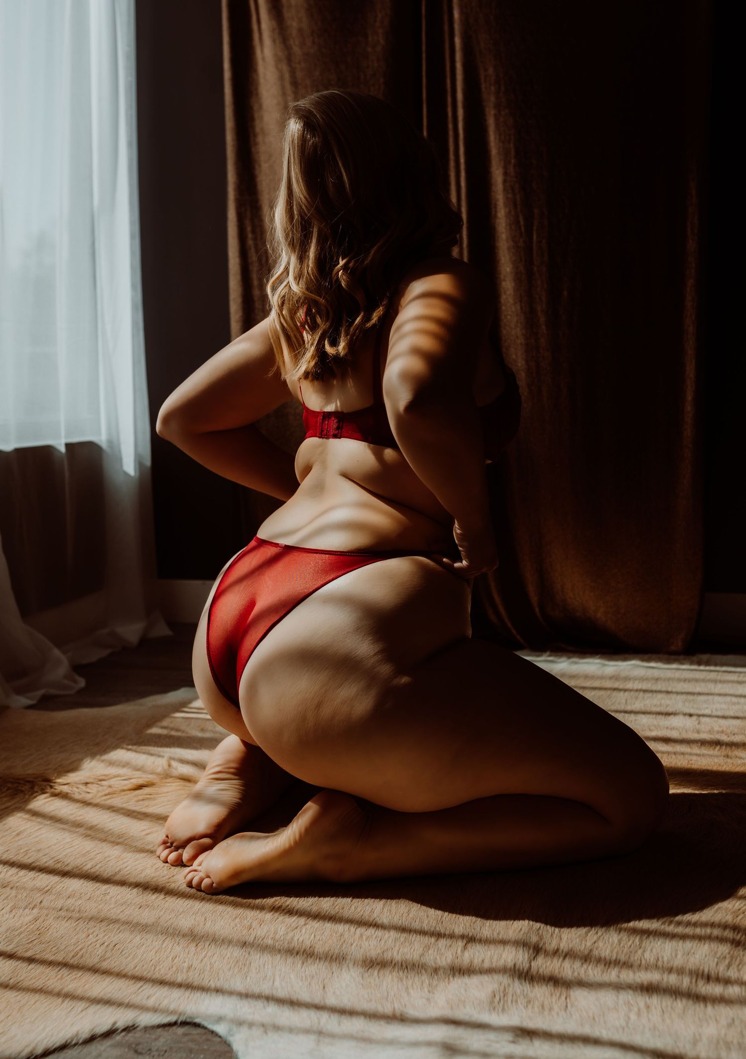 Woman in red bikini kneeling on sunlit floor with dramatic shadows, viewed from behind near curtained window.