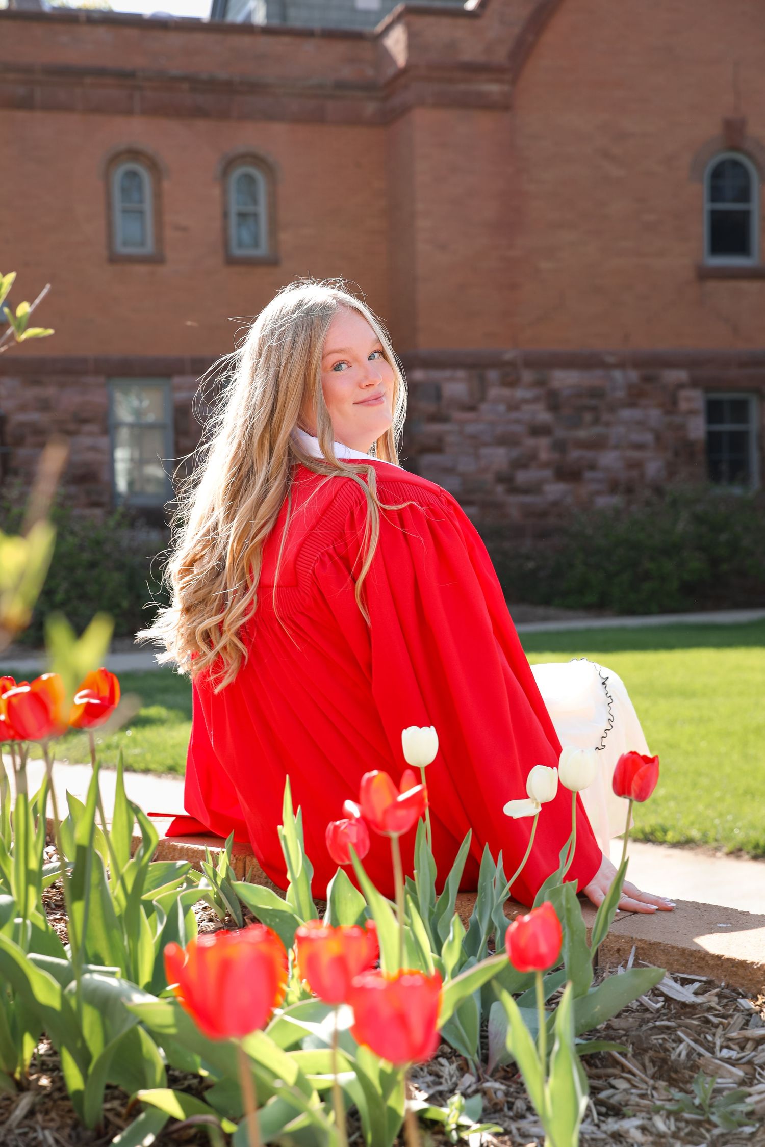 Young blonde girl in red graduation gown sitting among red and white tulips outside a brick building.