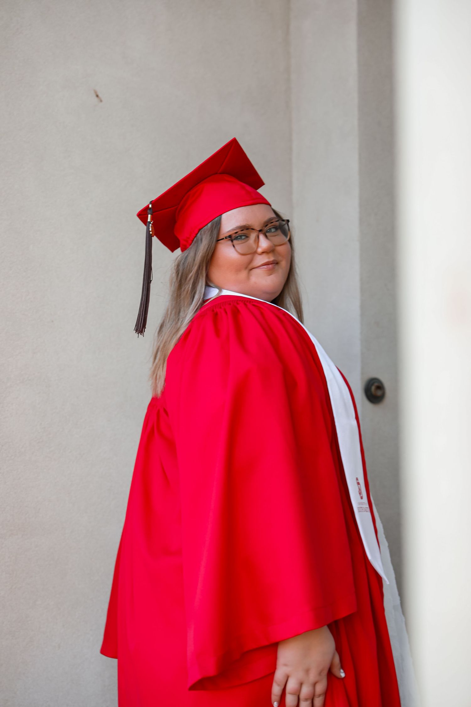 Young woman in red graduation cap and gown with glasses smiling over her shoulder outdoors.