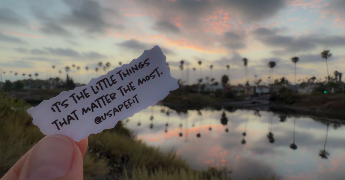 Hand holding a note saying 'It's the little things that matter the most' against a sunset sky over a tranquil lake.