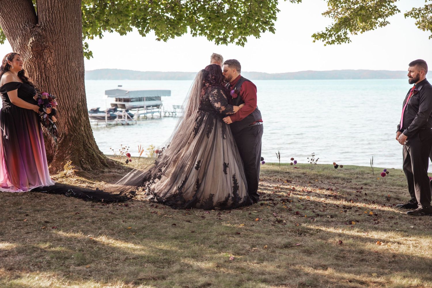 bride and groom first kiss under tree in front of Lake Michigan surrounded by bridesmaid and best man