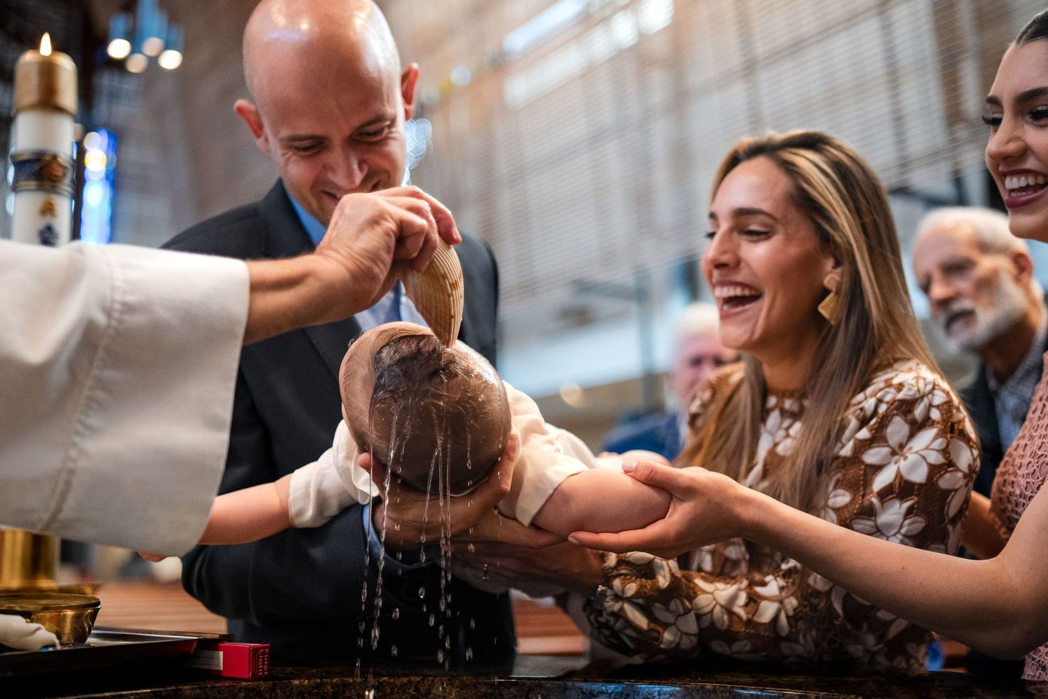 Parents smiling during a Catholic baptism ceremony as a priest pours holy water over an infant.