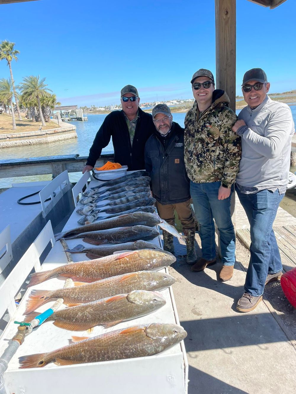 Group of four fishermen with large redfish and trout catch displayed on cleaning table.