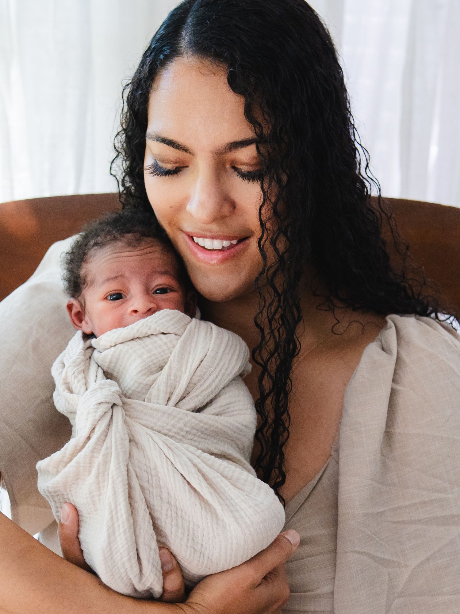A smiling mother with curly hair tenderly holds her swaddled newborn baby in a soft beige wrap.