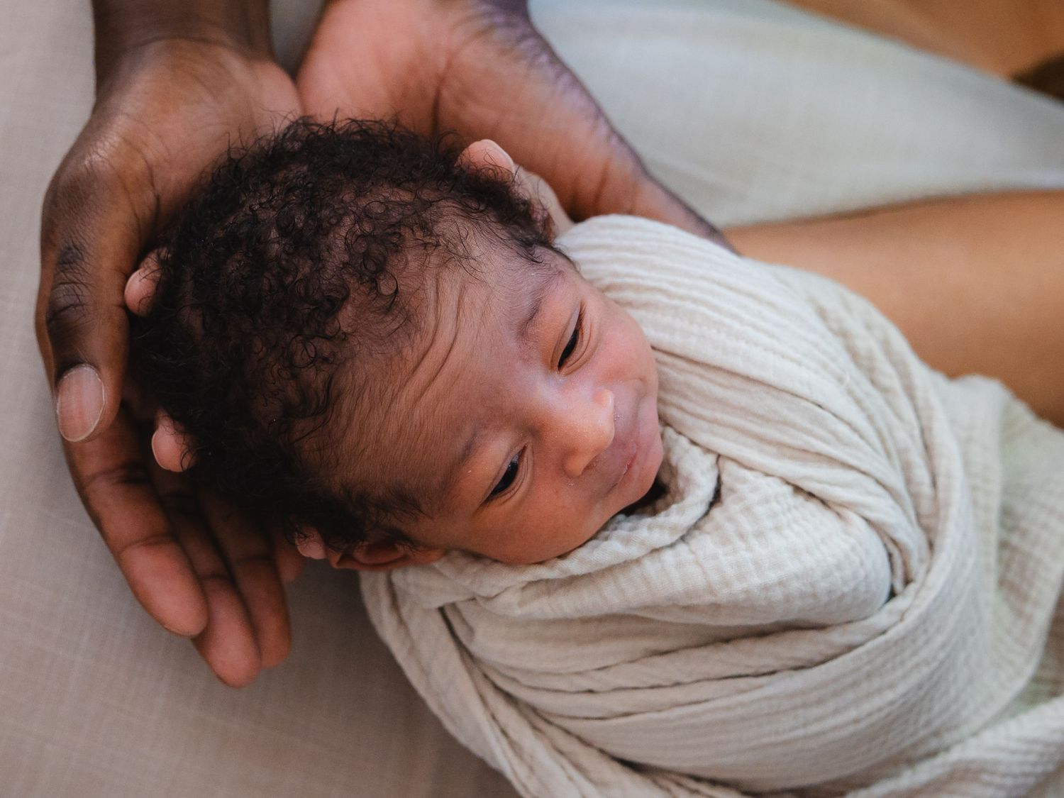 A newborn baby swaddled in a white muslin wrap rests peacefully in a parent's gentle hands.
