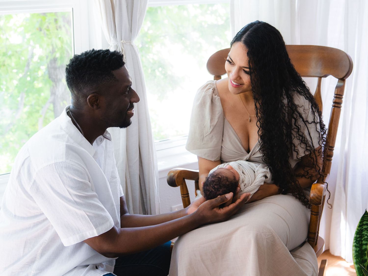 A couple tenderly gazes at their newborn baby, with the mother seated in a rocking chair near a bright window.