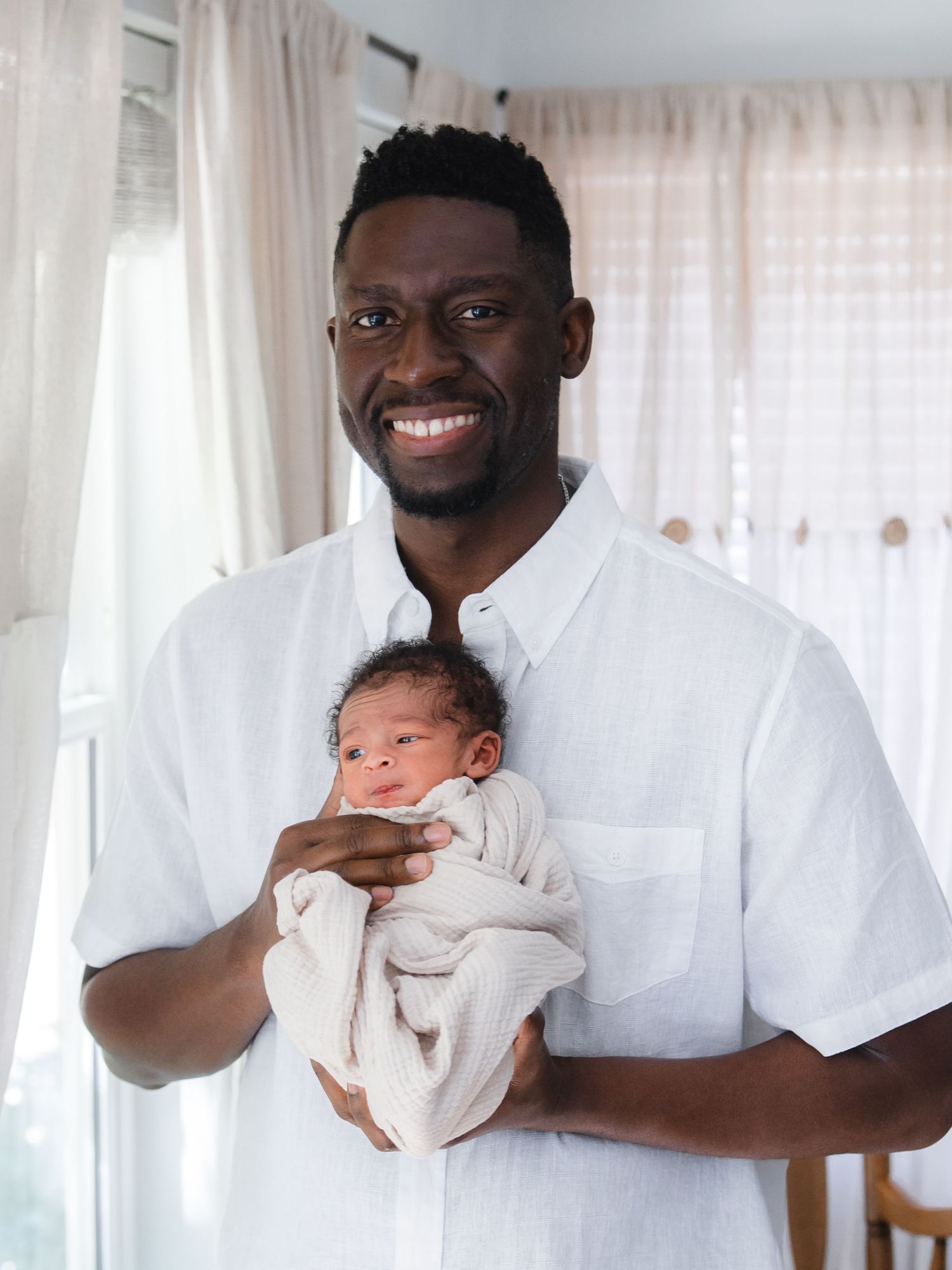 Smiling man in white shirt proudly holds a swaddled newborn baby indoors near bright curtained windows.