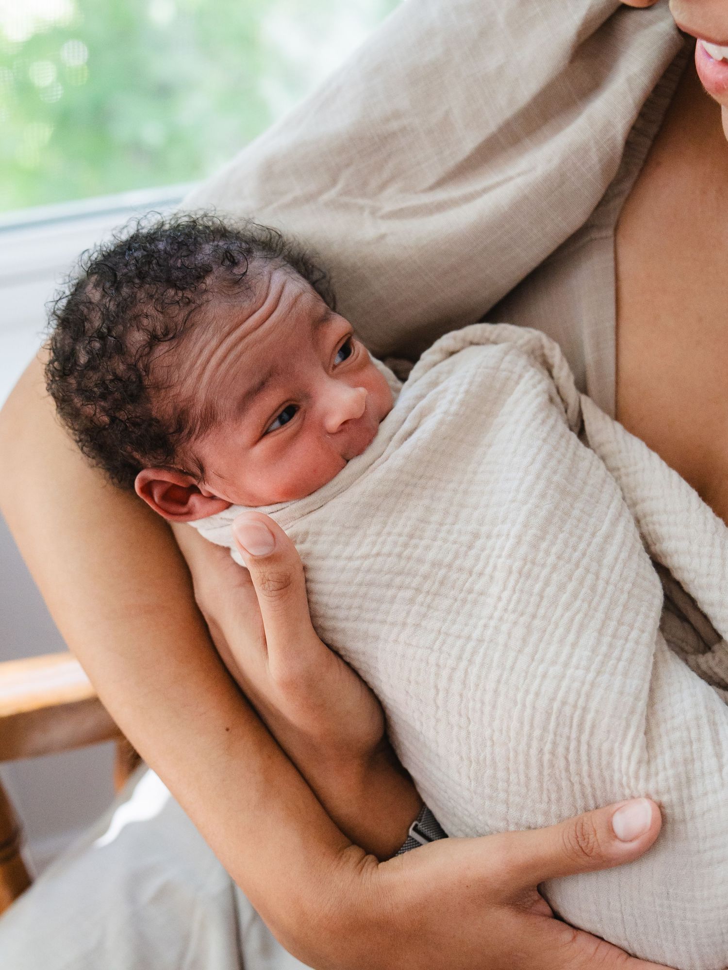 Newborn baby swaddled in cream muslin cloth, cradled in mother's arms near a bright window.