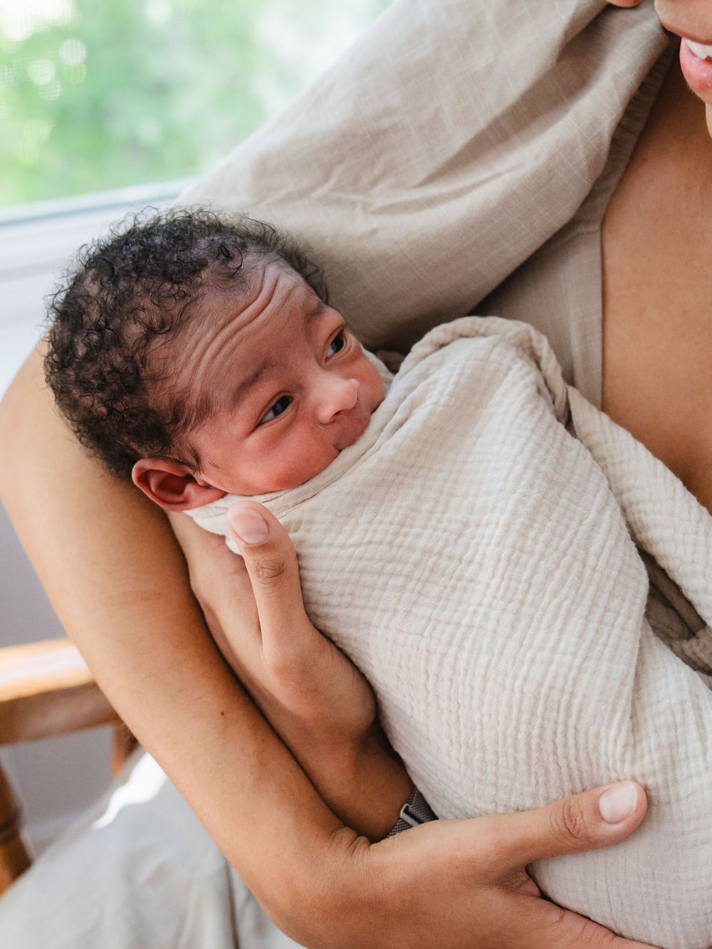 Newborn baby swaddled in cream muslin cloth, cradled in mother's arms near a bright window.