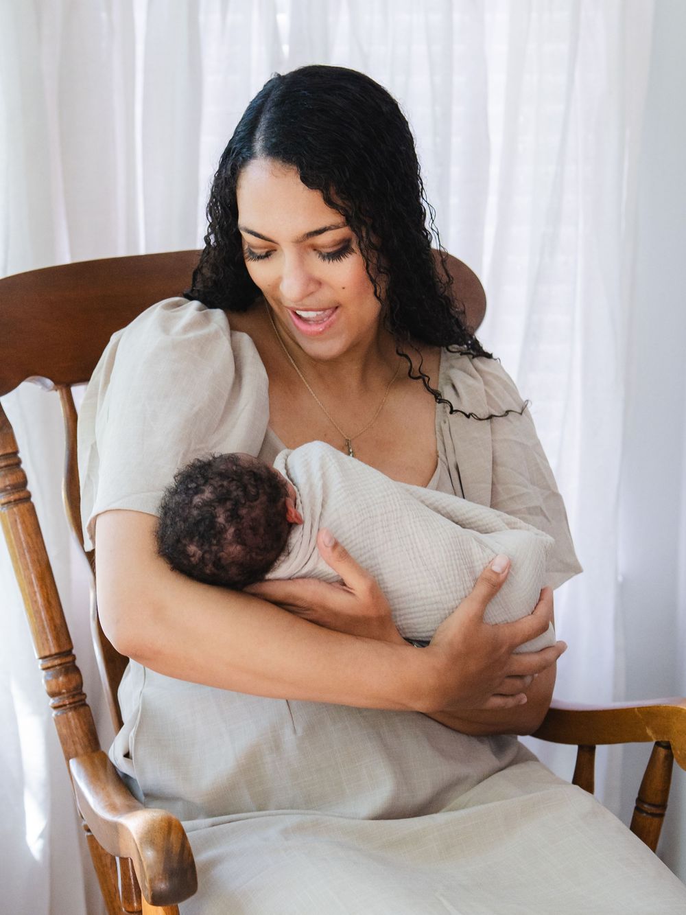 A mother sits in a wooden rocking chair, lovingly cradling her newborn baby wrapped in a white swaddle.