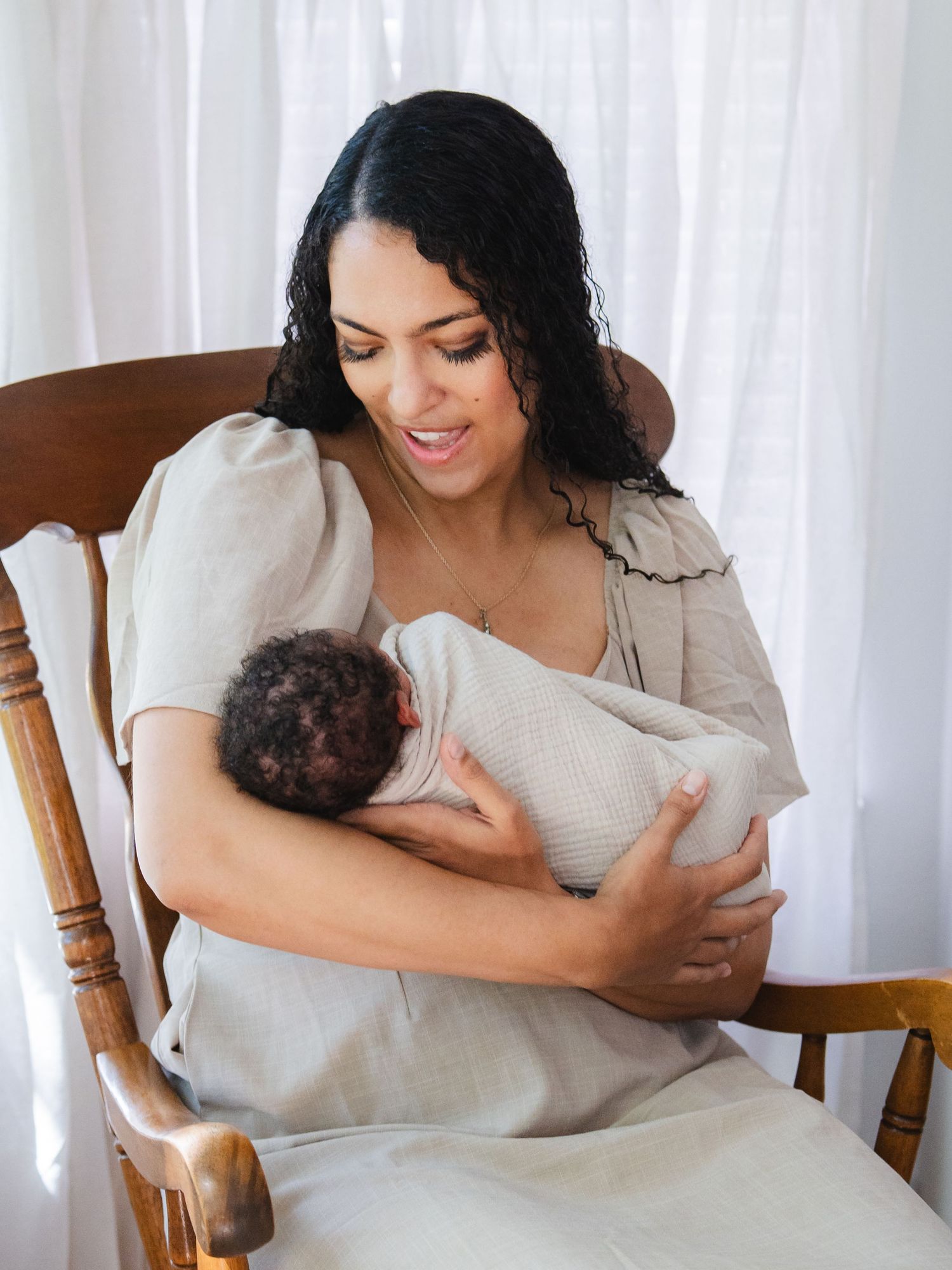 A mother sits in a wooden rocking chair, lovingly cradling her newborn baby wrapped in a white swaddle.