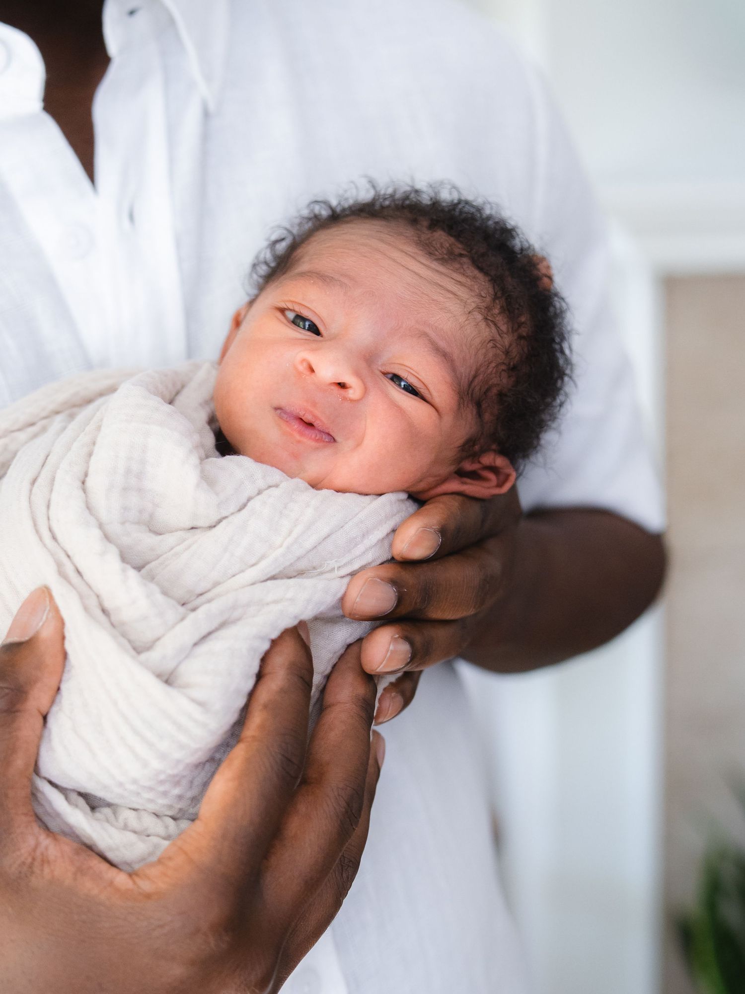 Newborn baby swaddled in white muslin cloth, cradled gently in adult hands, with soft natural lighting.