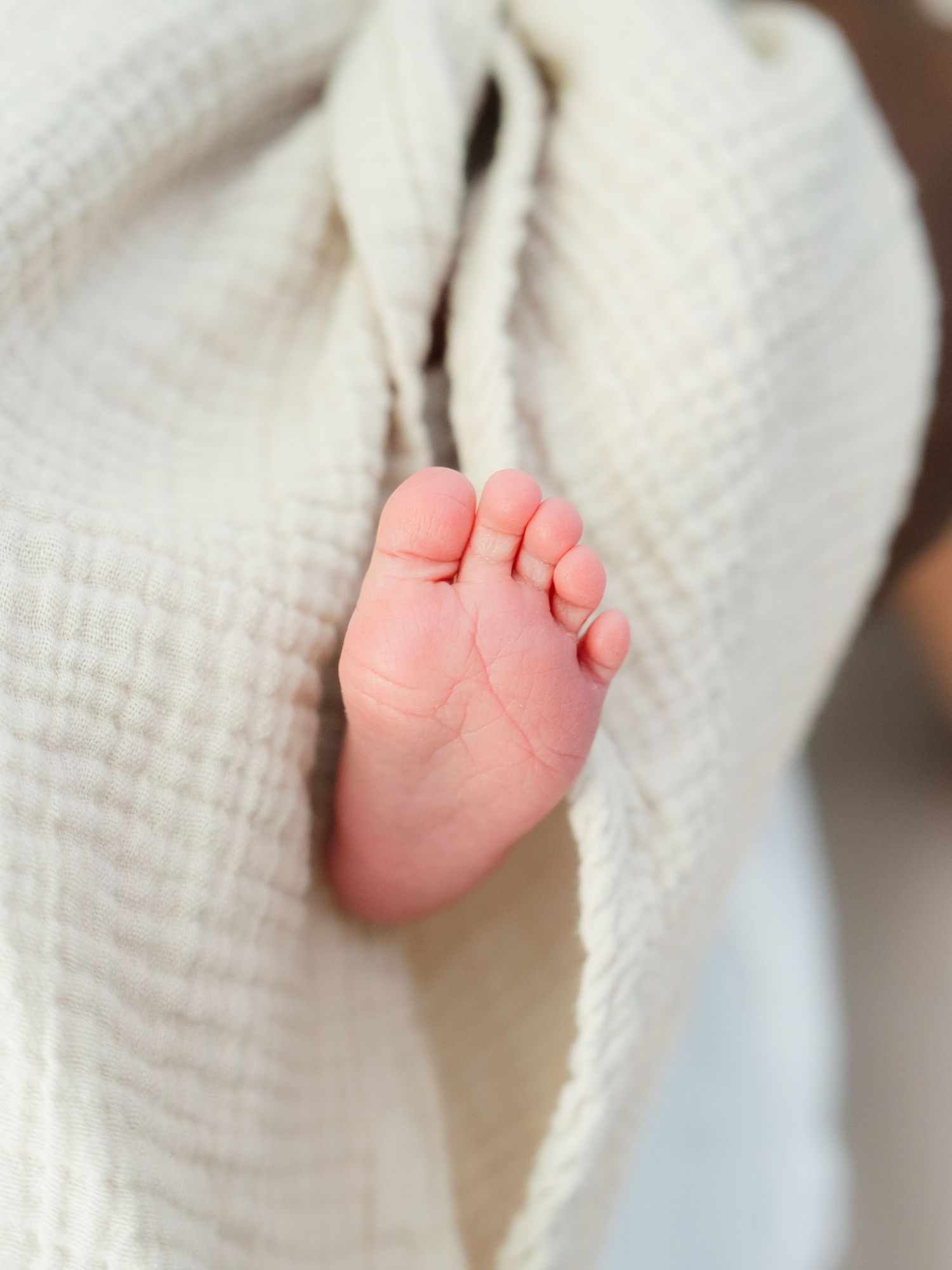 Close-up of a newborn baby's tiny foot peeking out from a soft white muslin swaddle blanket.