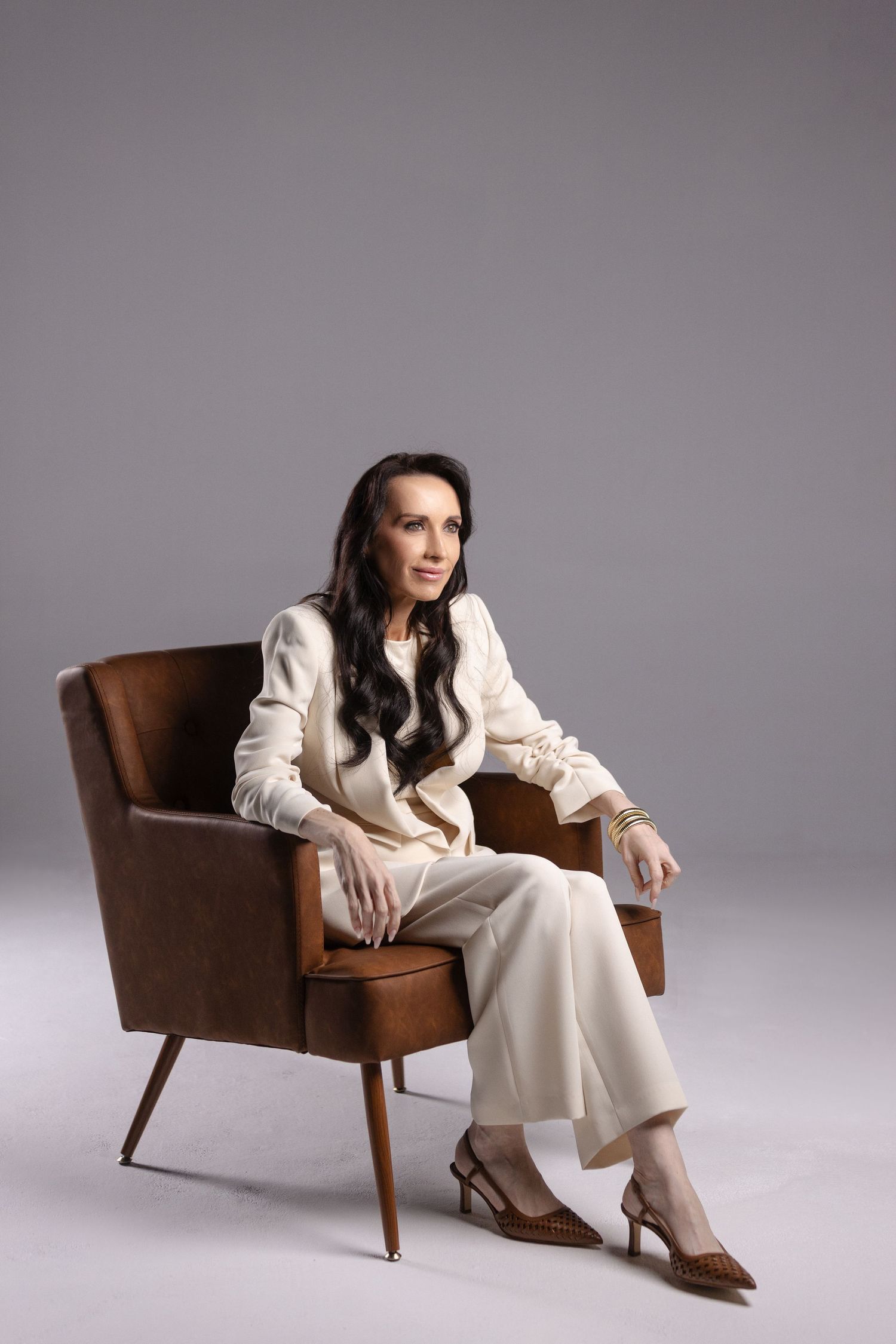 Blonde woman in white suit sitting on brown leather sofa in modern minimalist room with arched alcove and green plant.