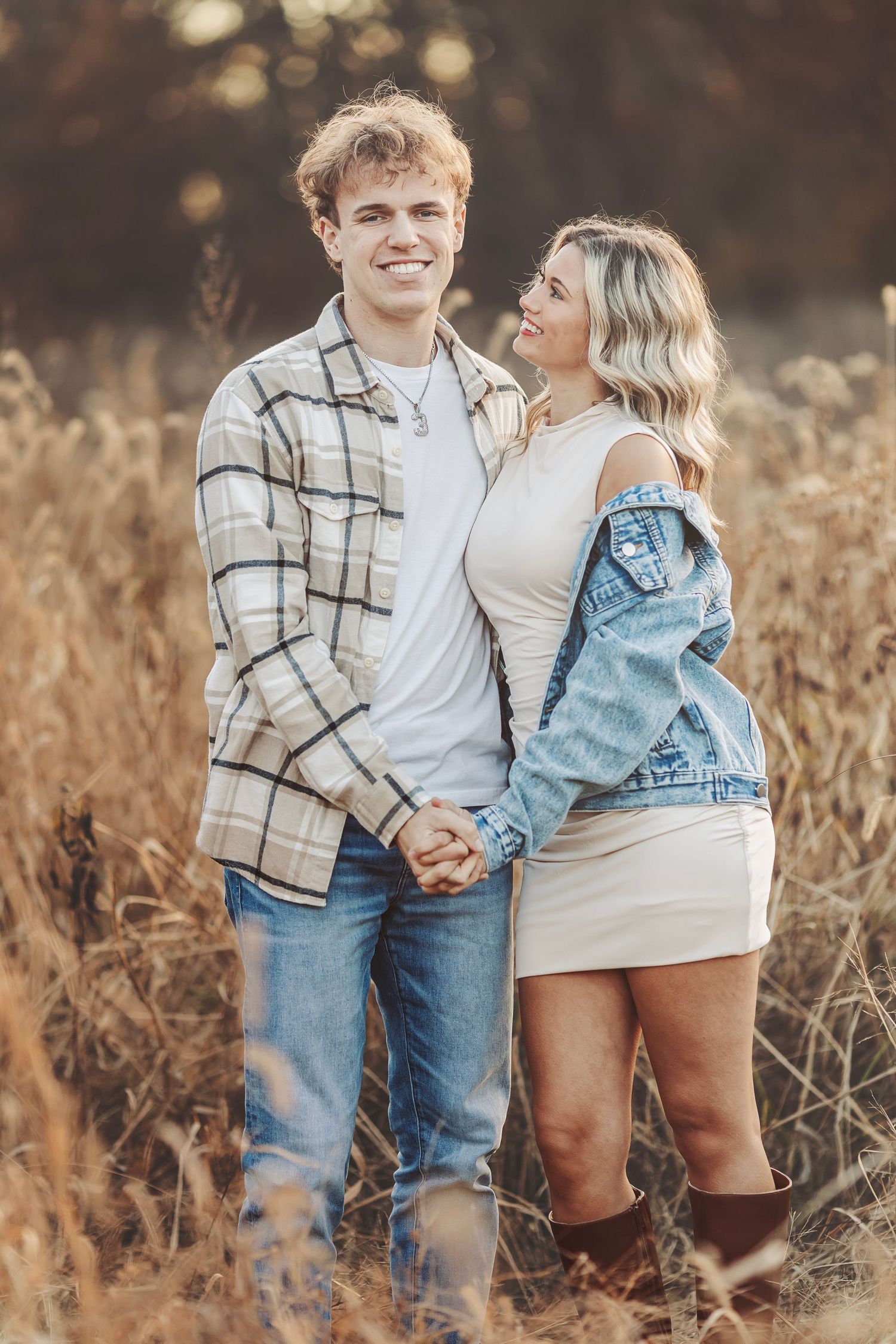 Couple poses outdoors in autumn park, woman in white sweater cradling baby bump, man in burgundy top embracing her from behind.