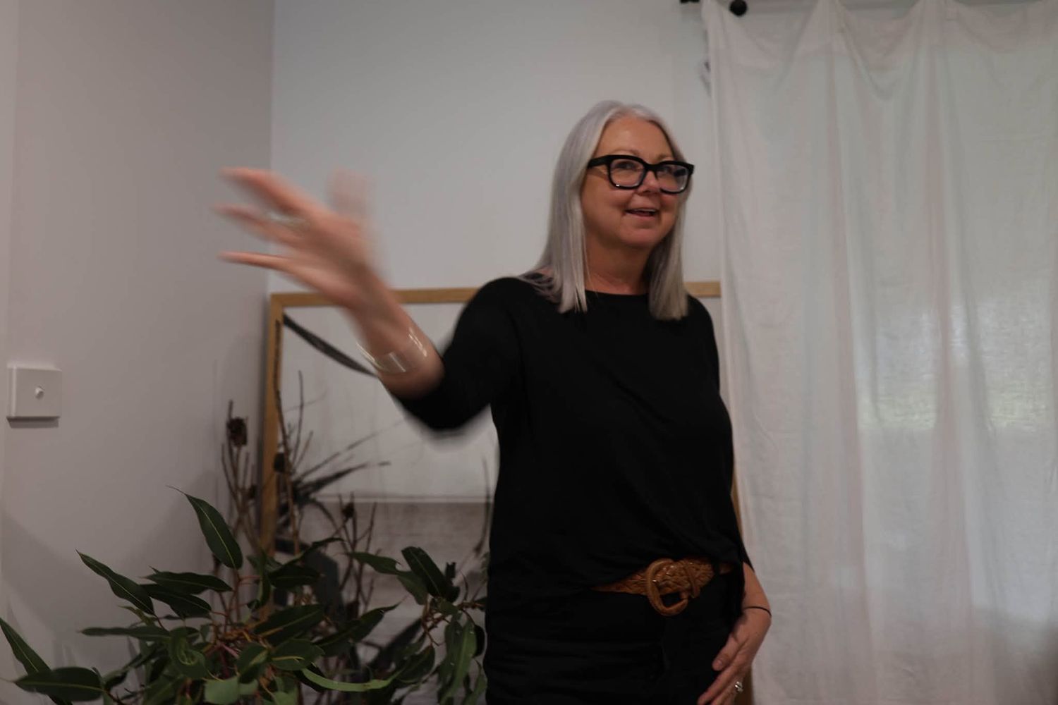 Woman in black outfit performing a high kick indoors near sheer white curtains and glass doors.