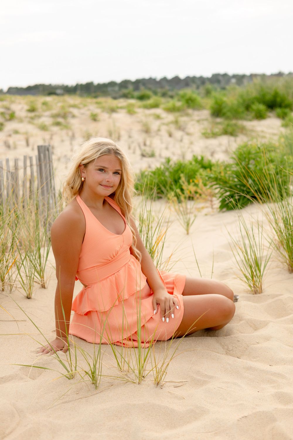 Young blonde woman in peach dress sitting on sandy beach dunes with grass and wooden fence backdrop.