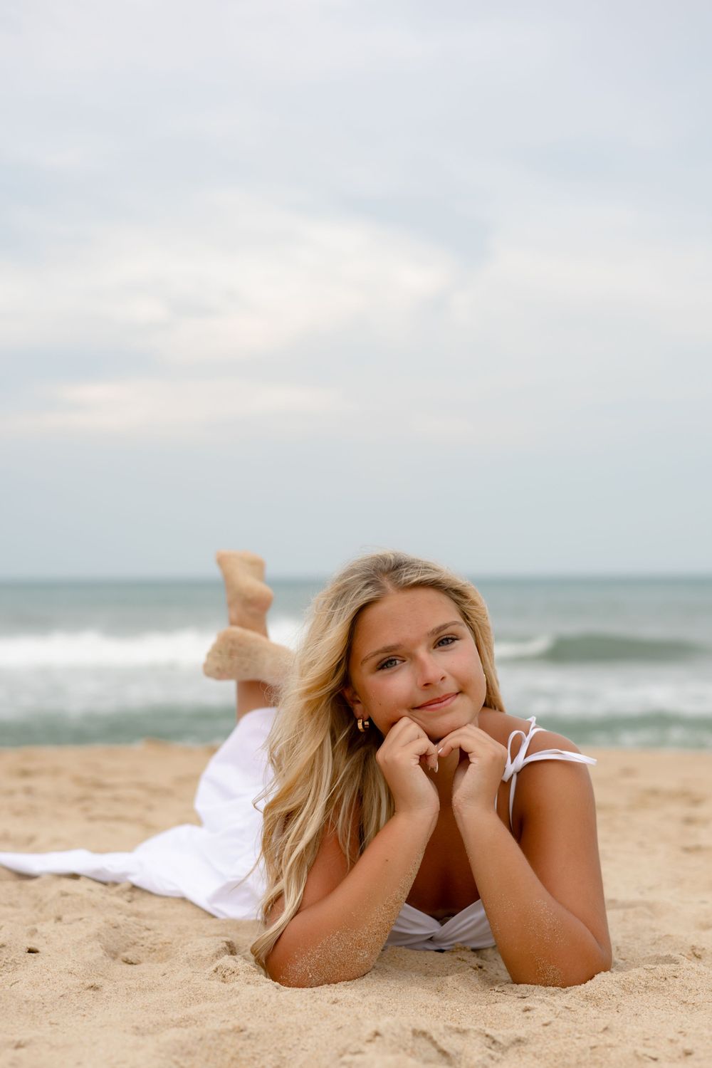 Young blonde woman in white dress lying on sandy beach with ocean waves in background on cloudy day.