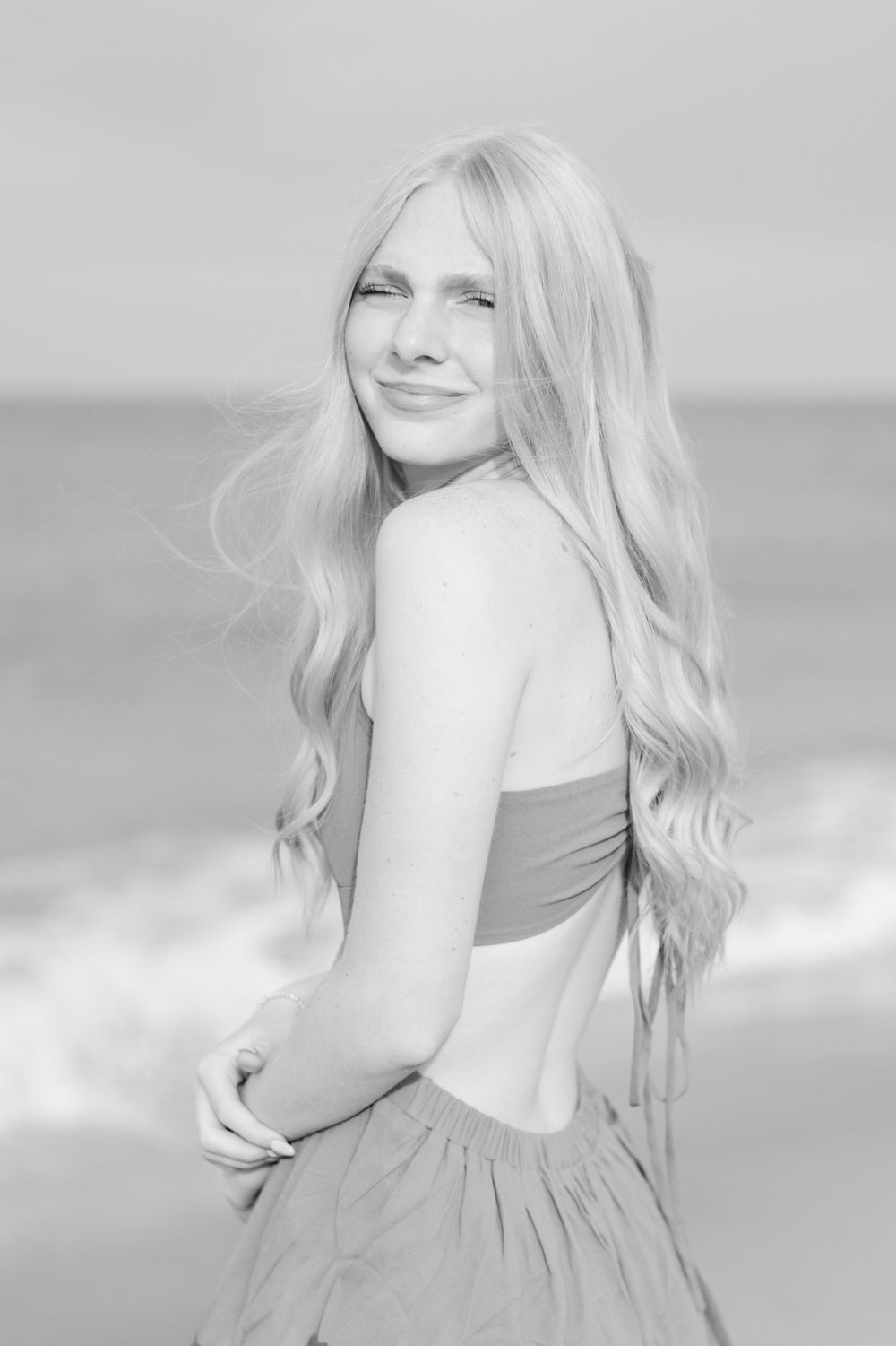 Black and white portrait of a blonde woman in a strapless dress smiling at the beach with ocean waves behind her.