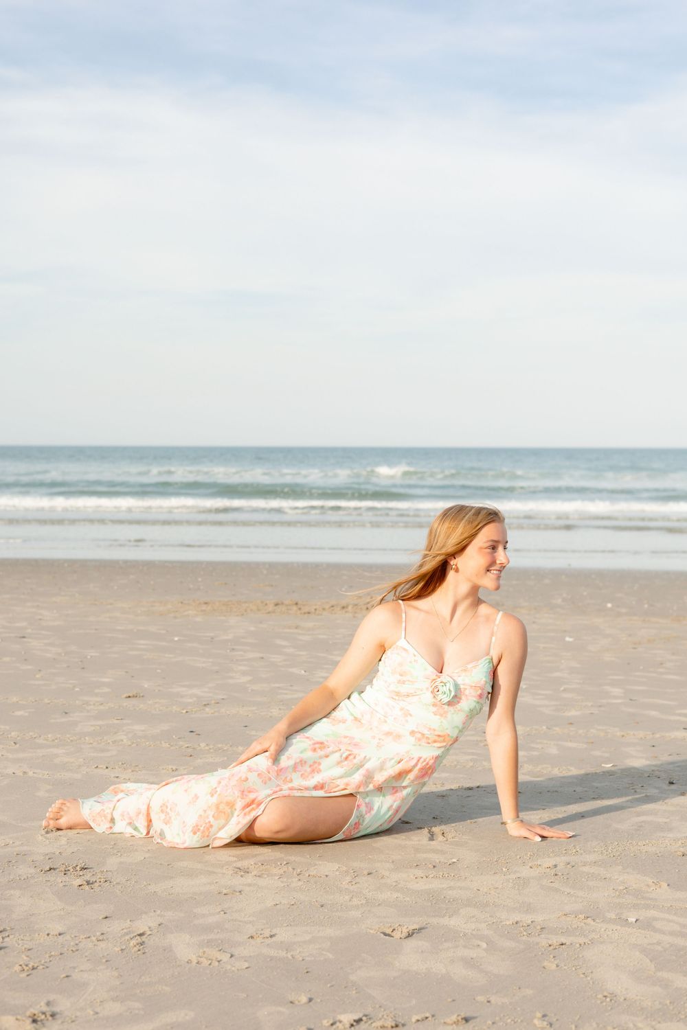 Young woman in floral maxi dress sitting on sandy beach with ocean waves in background on cloudy day.