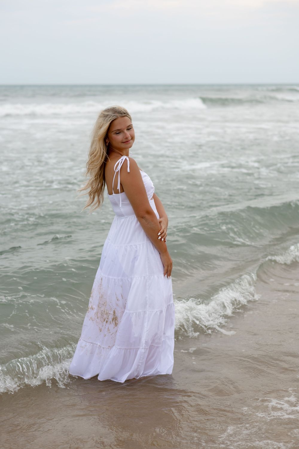 Blonde woman in white maxi dress standing in ocean waves on a beach, looking back over her shoulder.