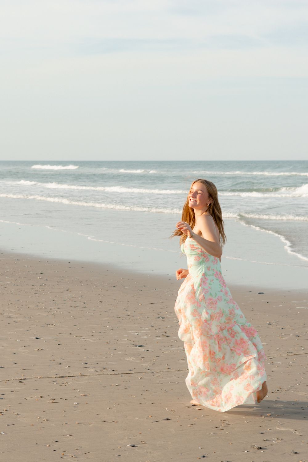 Young woman in floral maxi dress walking on sandy beach with ocean waves in background.