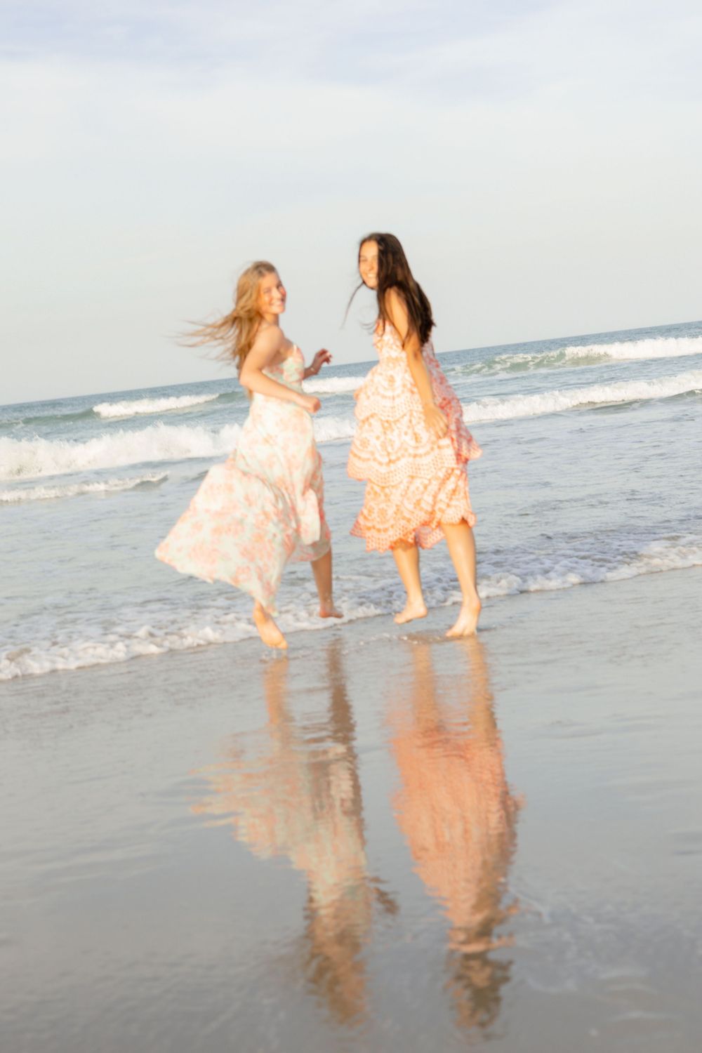 Two women in floral dresses dancing barefoot on a wet beach with ocean waves and their reflections visible.