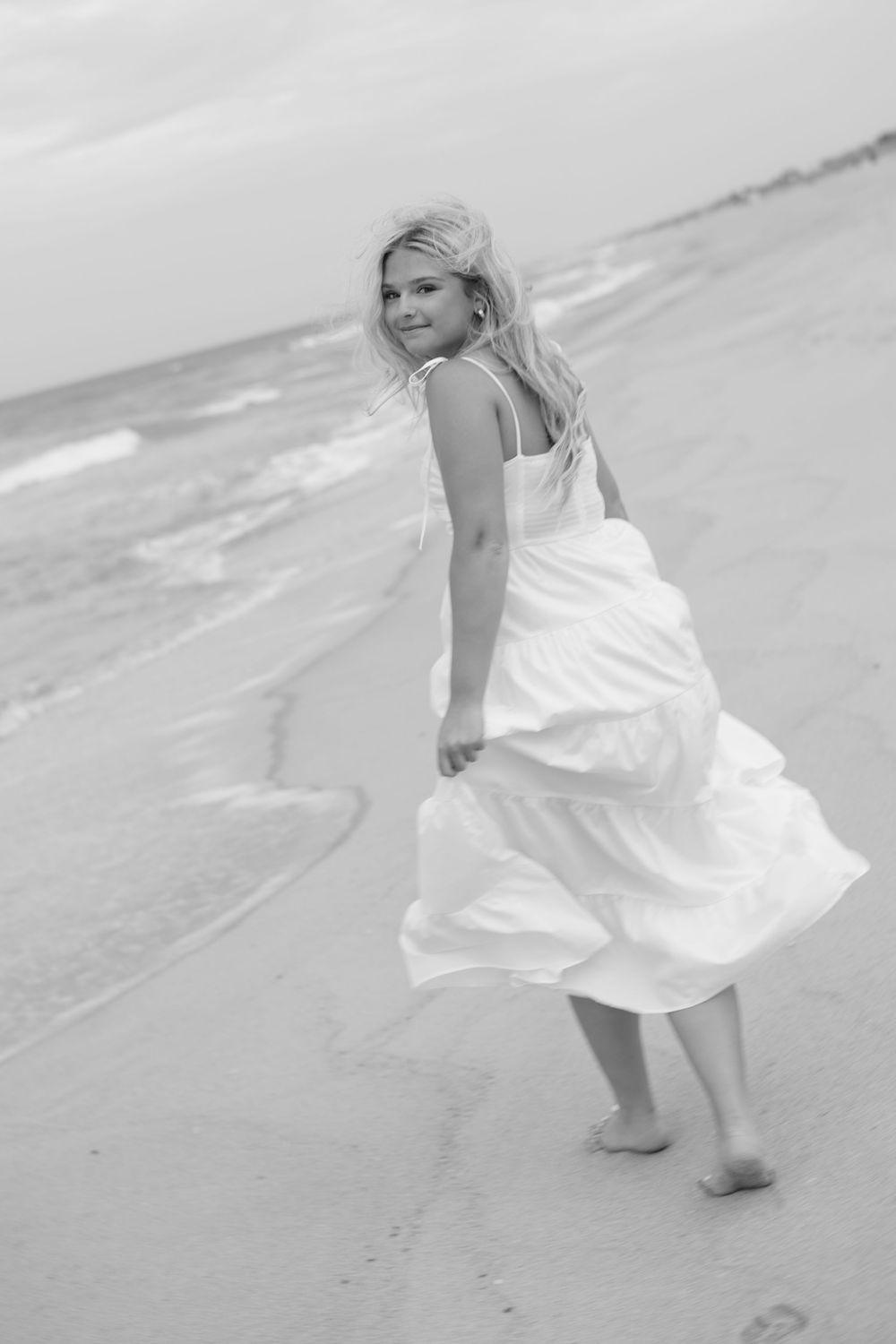 Blonde woman in white dress walking barefoot on beach, looking back and smiling, black and white photo.