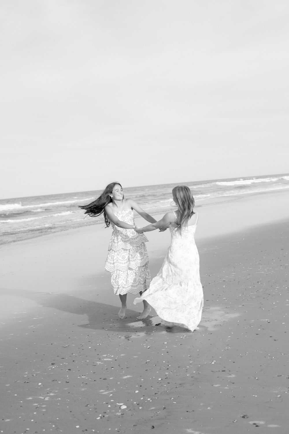 Two women in white dresses hold hands and dance joyfully on a sandy beach in a black and white photo.