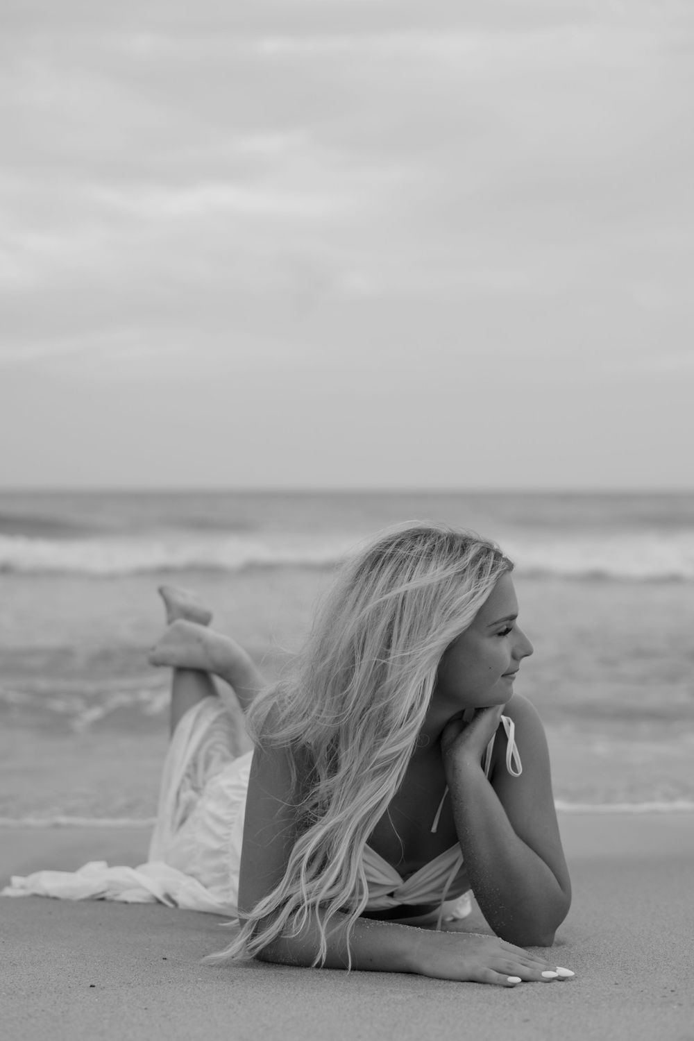 Black and white photo of a blonde woman lying on a beach, gazing at the ocean waves.