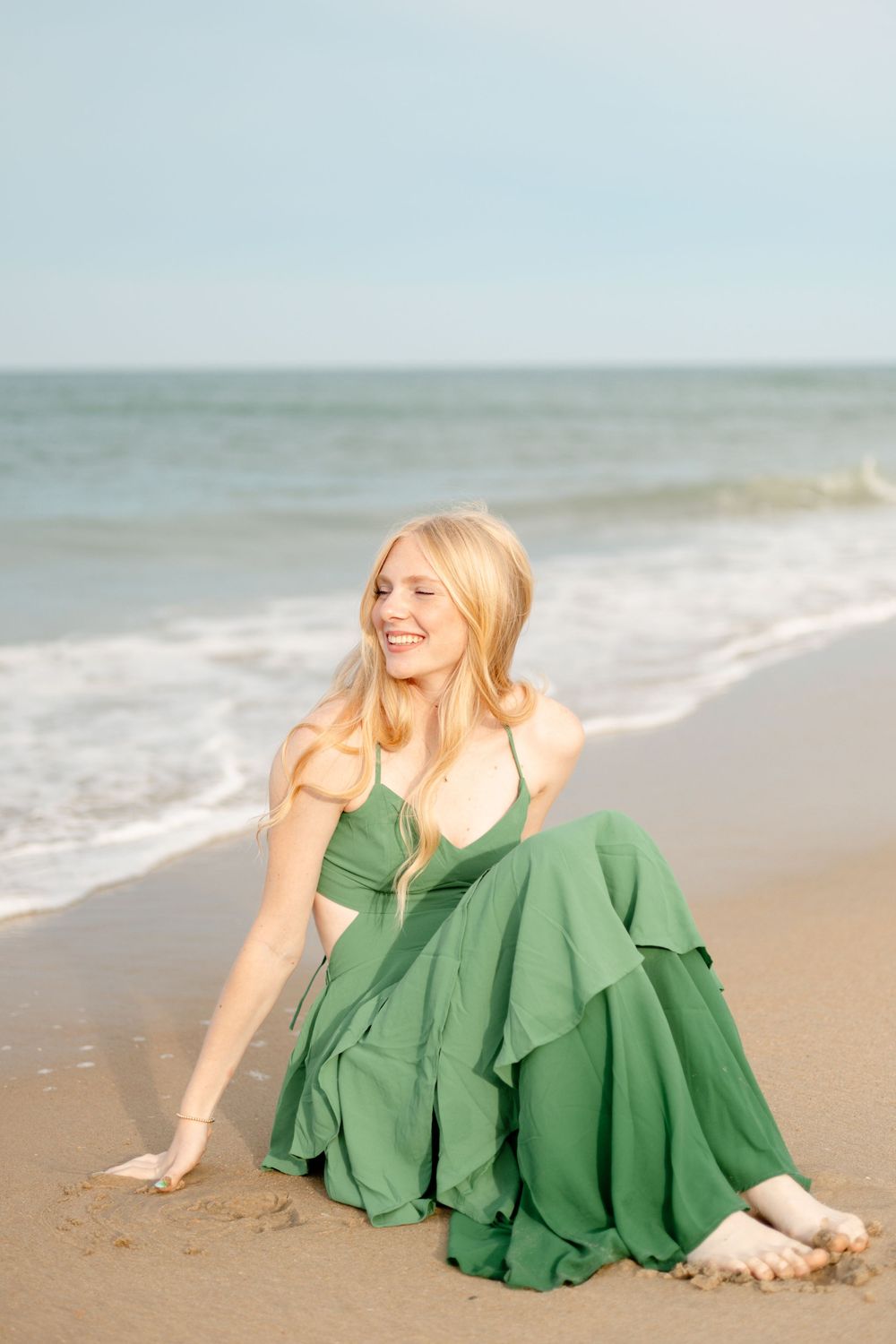 Smiling blonde woman in a green dress sitting on a sandy beach with ocean waves in the background.