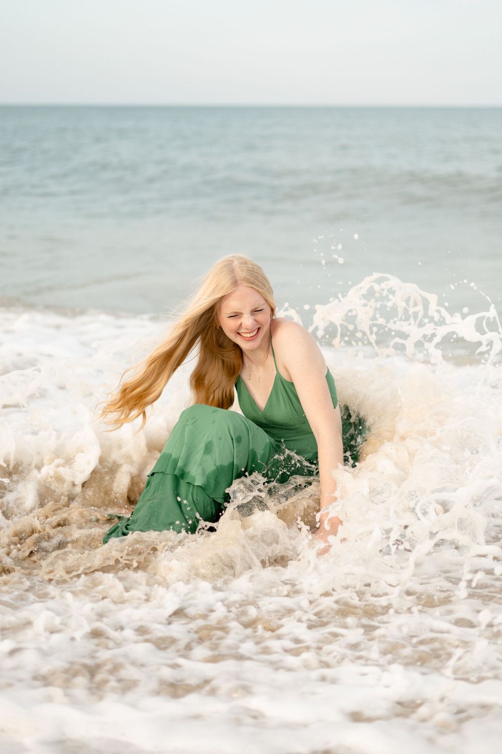 Smiling blonde woman in green dress sitting in ocean waves on beach with water splashing around her.