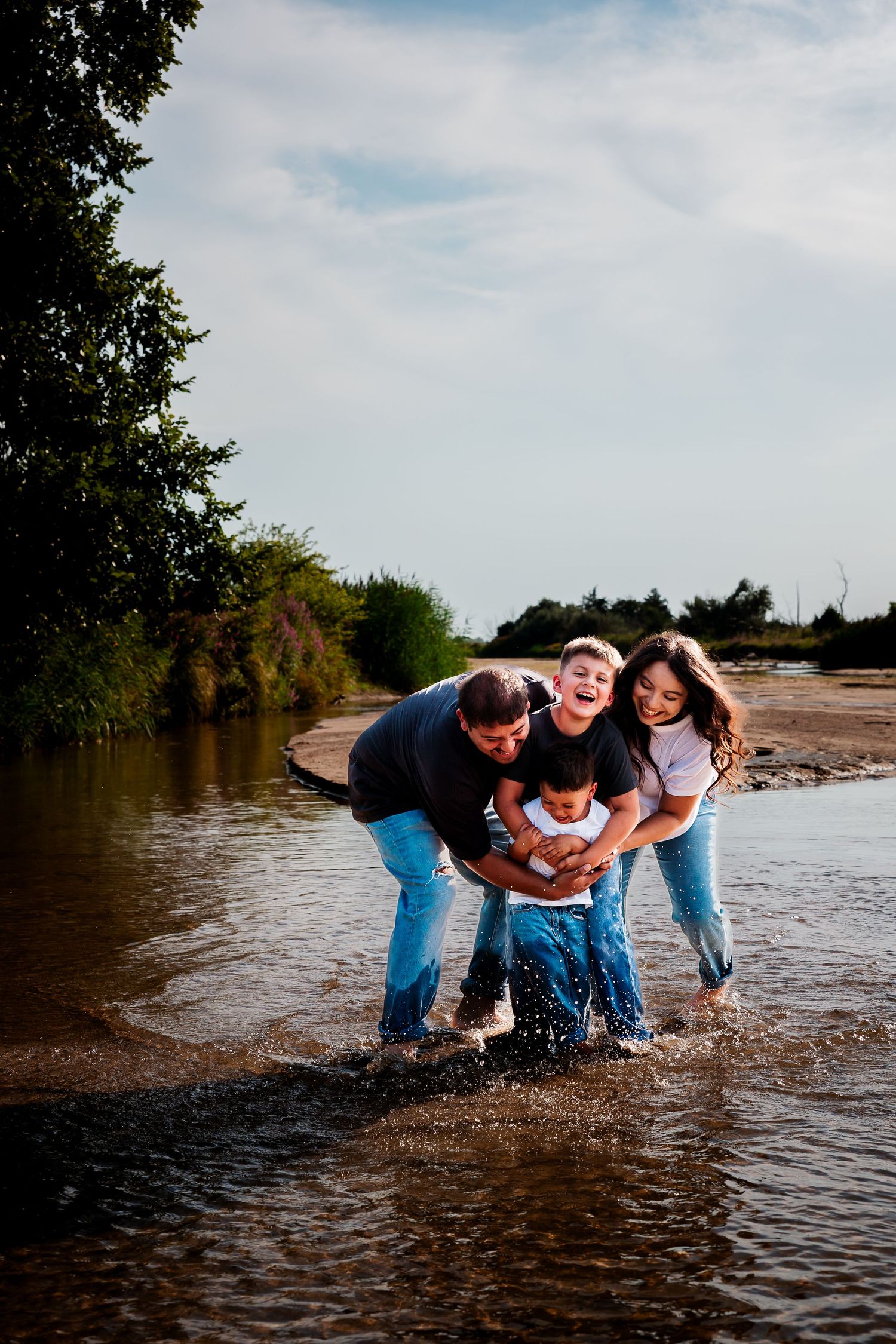 Family of four sitting together on a sandy beach, smiling and embracing with lush green trees in the background.