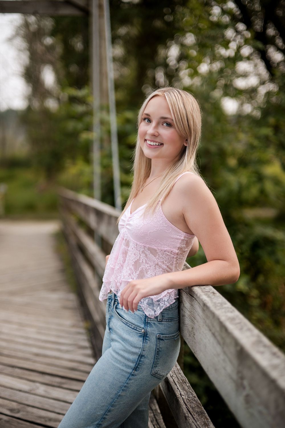 Smiling blonde teen girl in pink lace top and jeans leaning on wooden bridge railing outdoors.