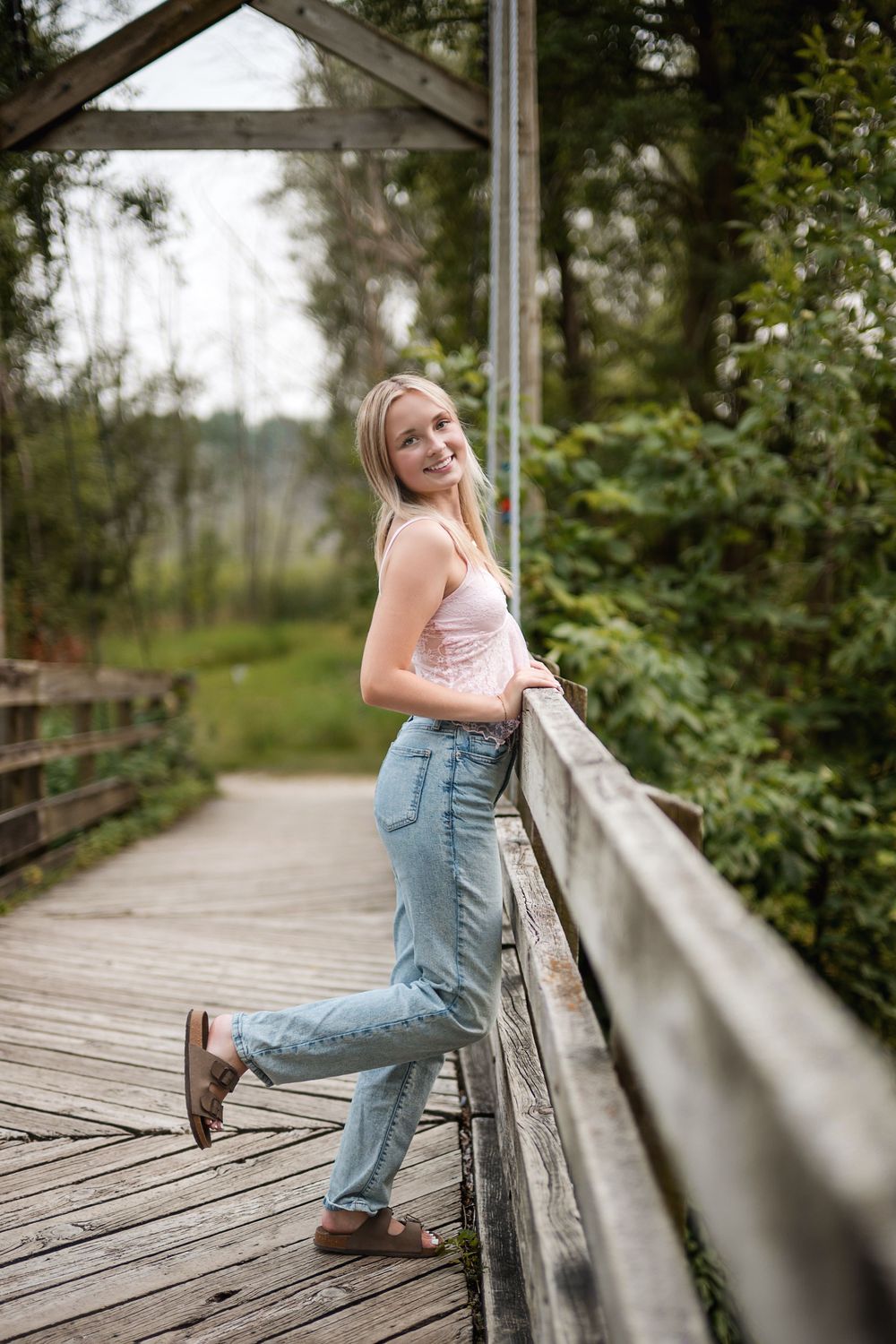 Young blonde woman in jeans and tank top leaning on wooden bridge railing in lush green park setting.