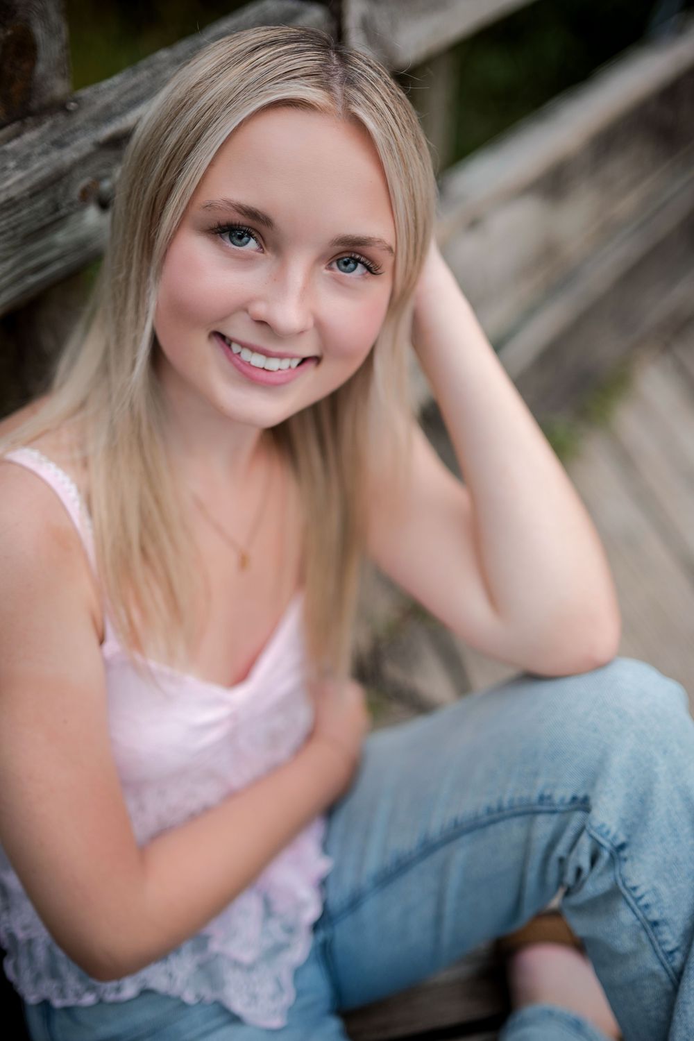 Pretty blonde teenage girl with blue eyes smiling in pink top and jeans sitting on wooden steps.