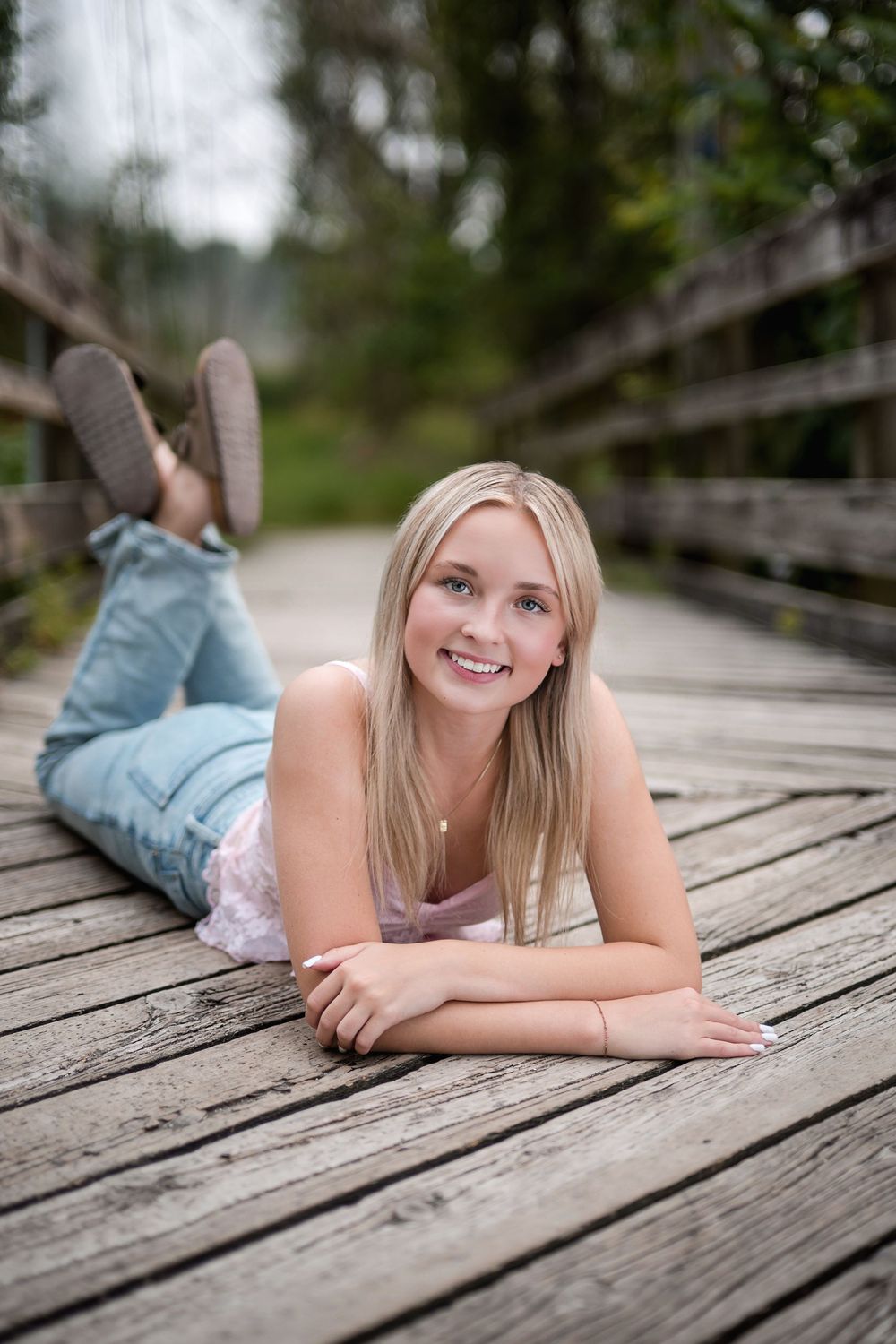 Smiling blonde teen girl lying on a wooden bridge boardwalk wearing jeans and a pink top outdoors.