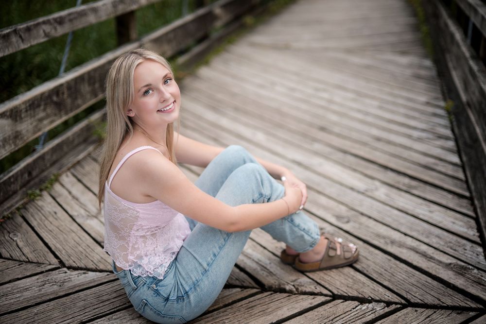 Smiling blonde teenage girl sitting on wooden bridge boardwalk wearing pink top and light blue jeans.