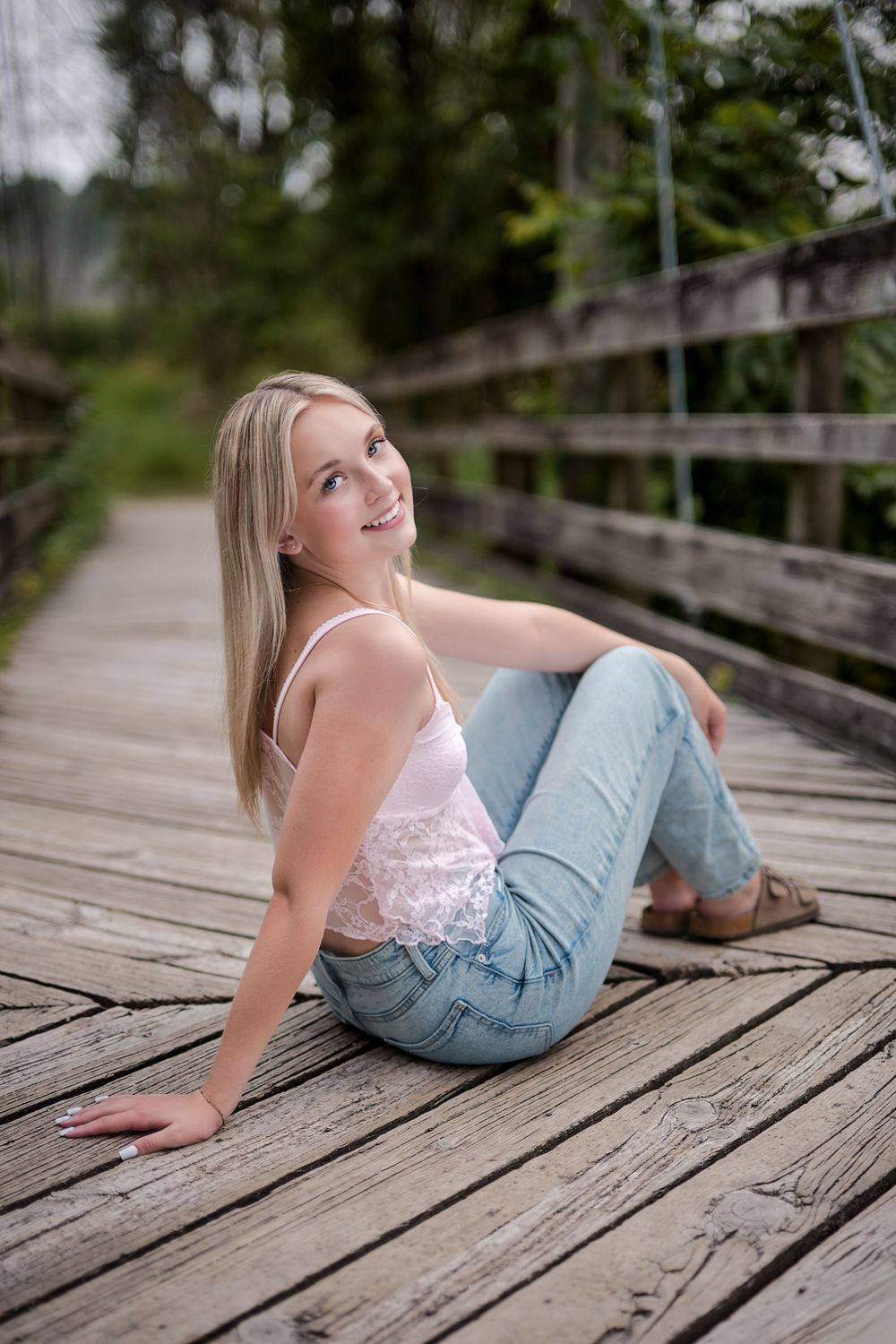 Smiling blonde woman in jeans and pink top sitting on a wooden bridge surrounded by lush greenery.