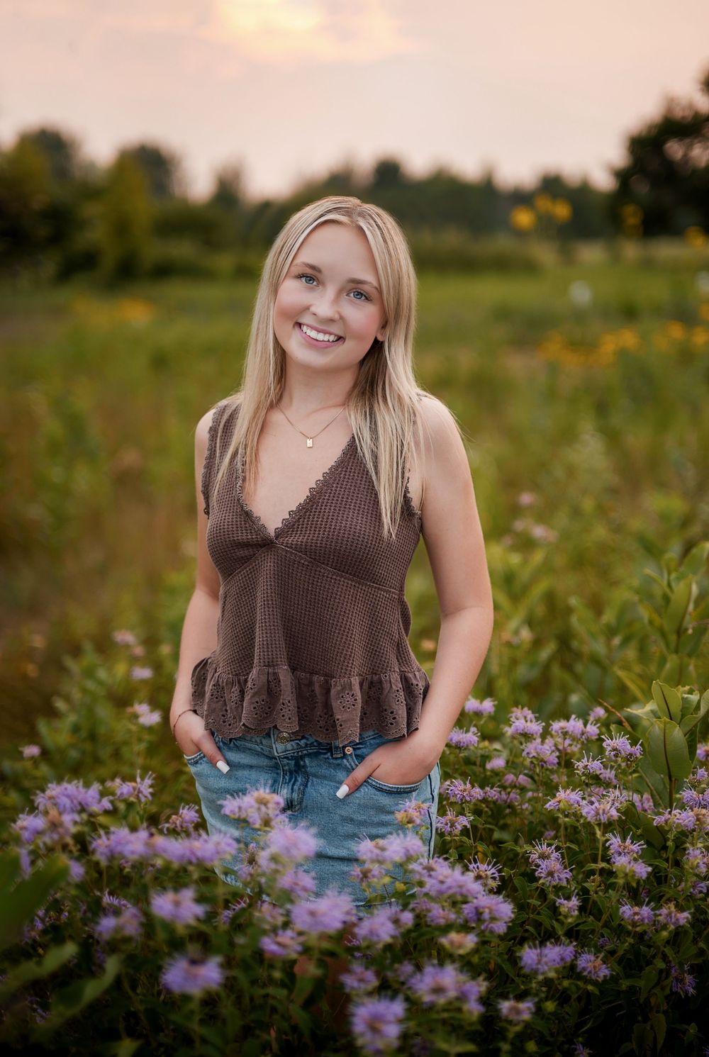 Smiling blonde woman in brown top and jeans standing among purple wildflowers in a sunlit meadow at golden hour.