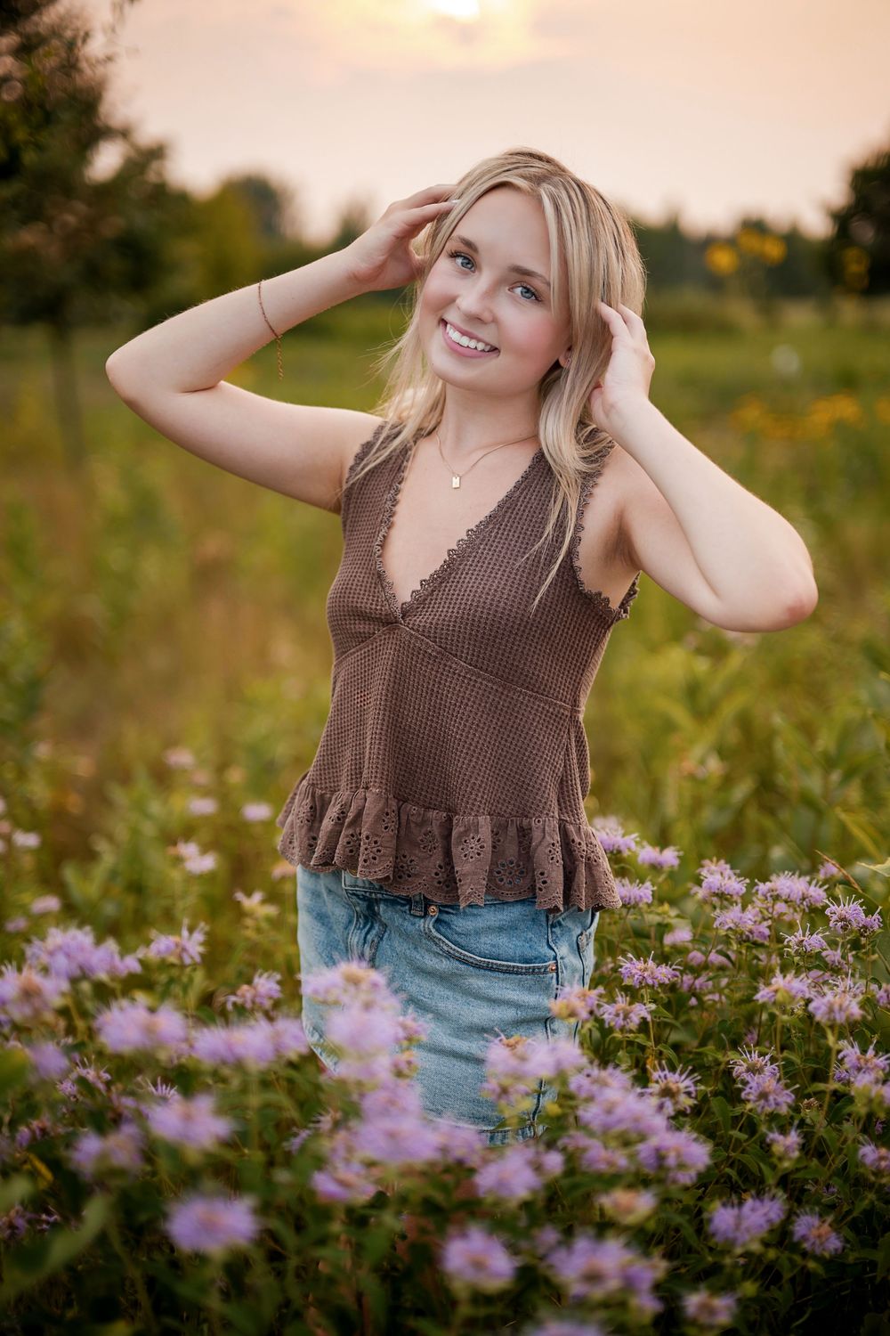 Smiling blonde woman in brown top and denim skirt posing among purple wildflowers at sunset.