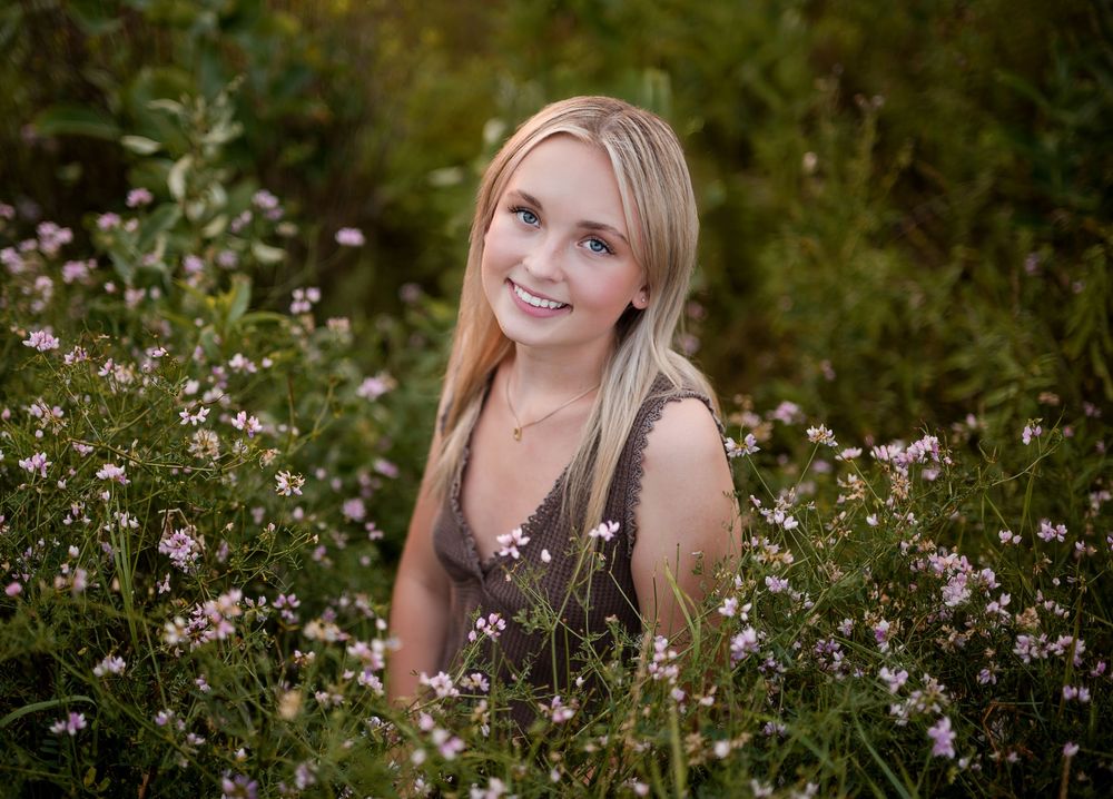 Smiling blonde woman surrounded by pink wildflowers in a lush green field, perfect for senior portrait photography.