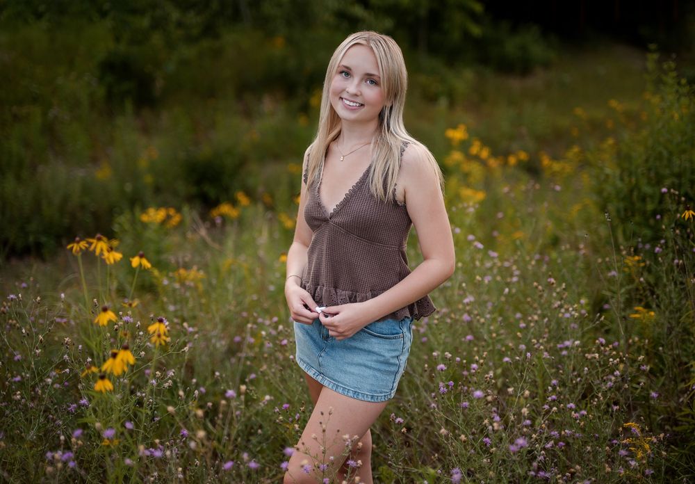 Young blonde woman smiling in a wildflower meadow, wearing a brown tank top and denim skirt.