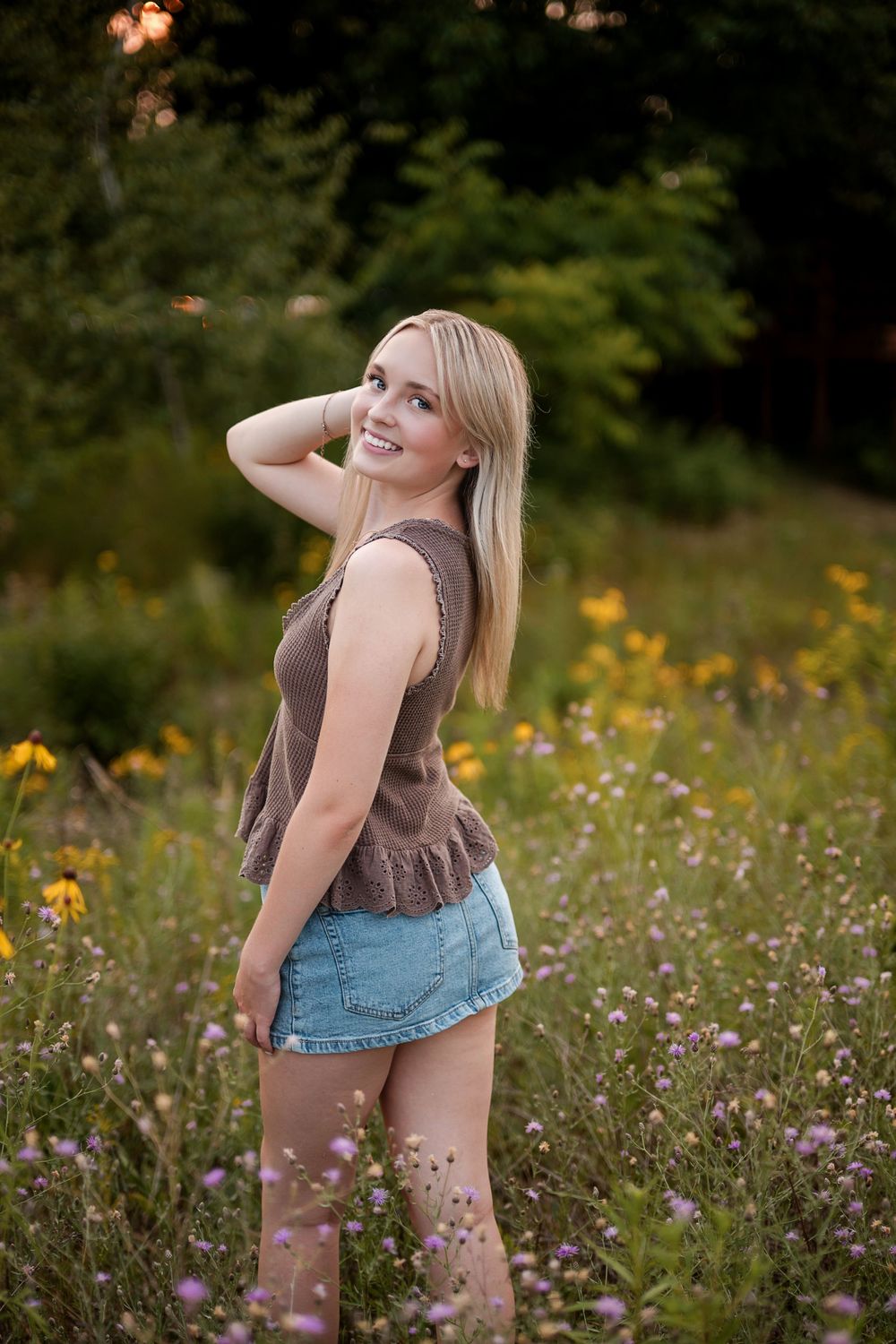 Blonde woman in brown peplum top and denim skirt smiling over shoulder in colorful wildflower field.