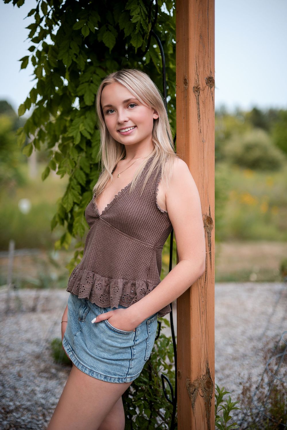Smiling blonde teen in brown lace top and denim skirt posing by wooden post with green vines.