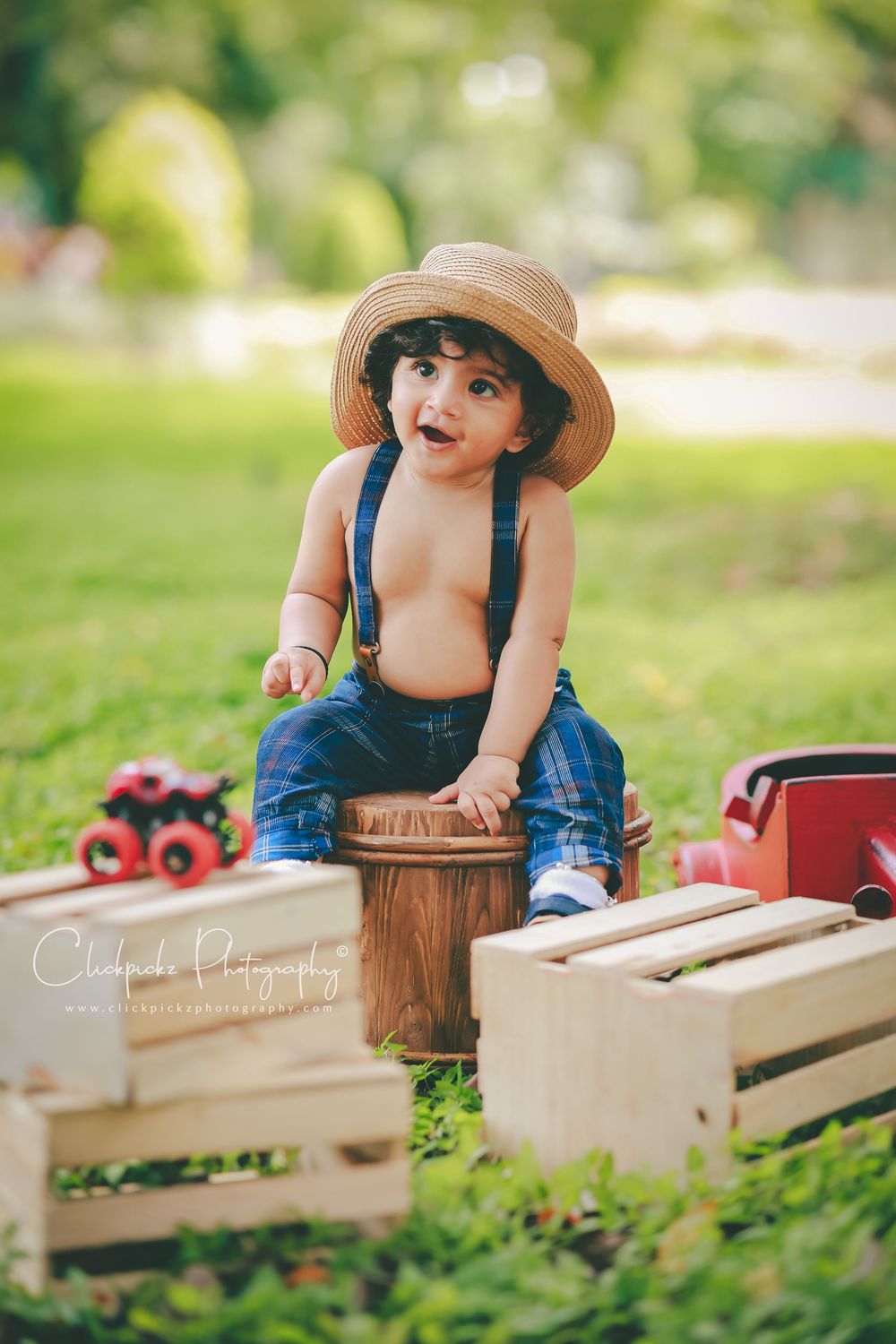 Toddler boy in straw hat and suspenders sits on wooden barrel outdoors among rustic crates and toy trucks.