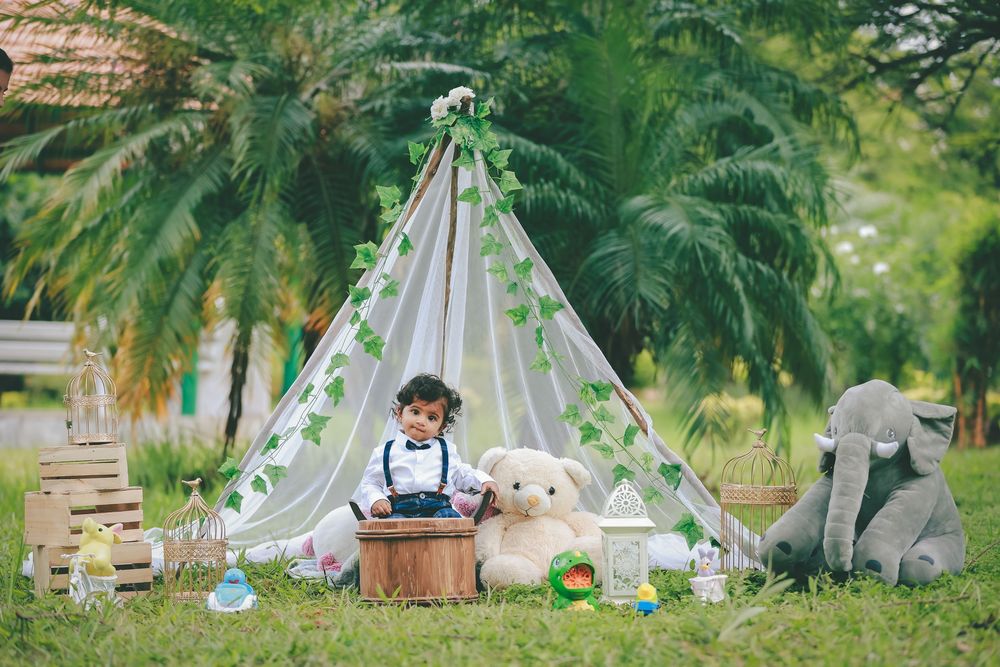 Toddler boy sits between a teddy bear and elephant plush toy inside a decorated teepee tent outdoors.