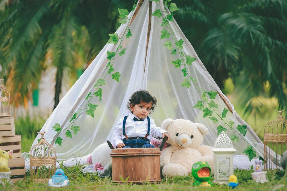 Young child sitting by a decorated teepee tent with a teddy bear and toys on green grass outdoors.